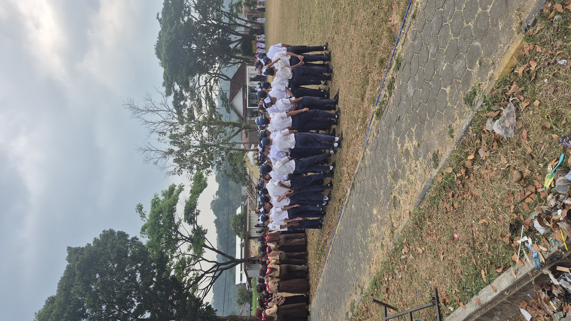 In Tambahsari, Indonesia, a group of Indonesian students, likely in uniform, are gathered in a semi-circle on a dry, grassy field. The students on the left are wearing brown uniforms with scarfs, resembling scouts. The students on the right are in white shirts and dark pants, possibly school uniforms. They are standing and facing inward, suggesting a briefing or assembly. In the background, several buildings with white walls and tiled roofs are visible, along with trees and a hilly landscape under an overcast sky. The foreground shows a paved pathway made of hexagonal stones, bordering the field. A concrete curb runs along the edge of the field, with dry leaves and some litter scattered on the grass and along the curb.