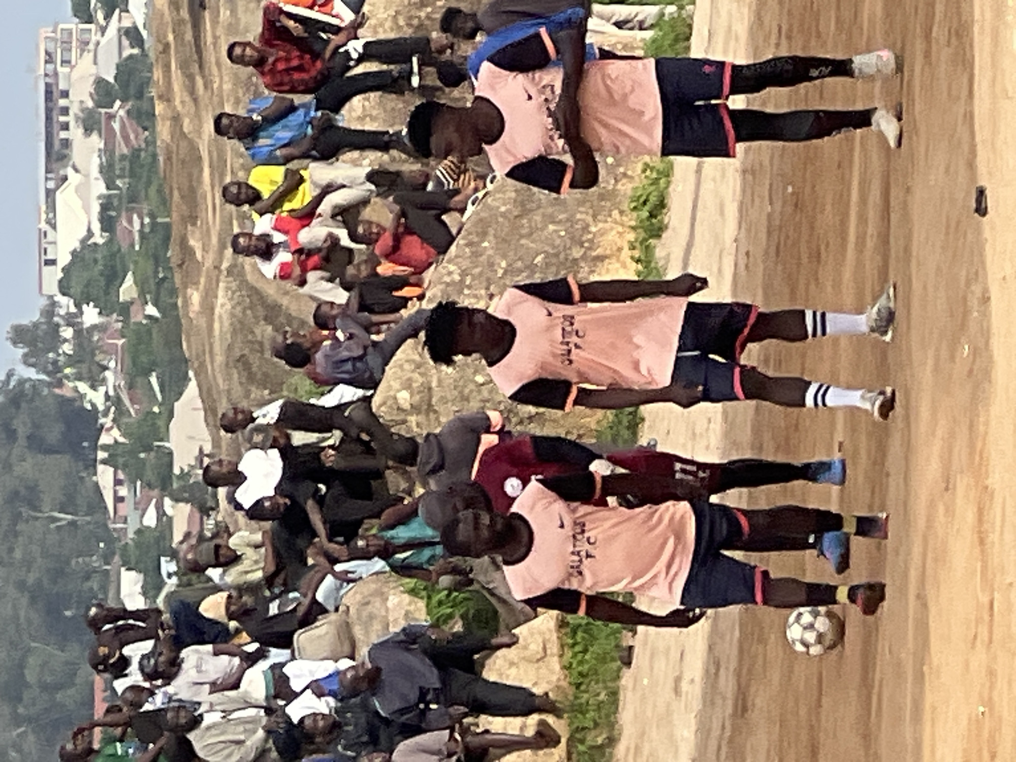 The image depicts a dusty outdoor scene, likely a sports field, with a group of young men in athletic attire. Three young men in the foreground are wearing pink and black soccer jerseys and dark shorts. Two of them are facing the camera, while one is partially turned away. A soccer ball is visible on the ground near the feet of one of the men. In the background, a crowd of people, appearing to be spectators, are seated or standing on a rocky incline. They are wearing a variety of casual clothing. Beyond the spectators, there are buildings and trees, suggesting an urban or semi-urban environment. The overall impression is a community gathering for a soccer match in a dusty, outdoor setting. The presence of the stadium and the rocky incline suggests the location might be a public area or a local sports facility within the city of Jos, Nigeria, a region known for its vibrant community sports culture.