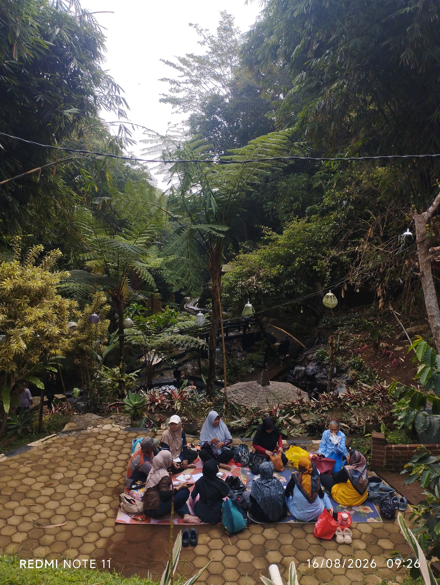 In Subang, Indonesia, a group of approximately ten individuals, predominantly women wearing hijabs, are gathered for a picnic on a patterned paved area surrounded by lush greenery. They are seated on colorful mats spread on the ground. Some are engaged in conversation, while others appear to be preparing or consuming food. Several bags, including backpacks and tote bags, are scattered around them, along with a few pairs of sandals. The background features a dense forest with bamboo and fern trees, as well as a small waterfall or stream. Decorative lamps are suspended from wires overhead, adding to the ambiance of the natural setting. The overall scene depicts a relaxed outdoor gathering in a verdant, possibly hilly, environment.