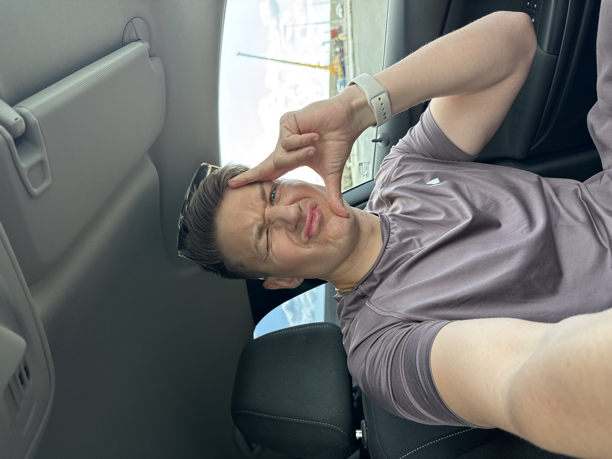 A medium-shot, selfie-style photograph taken inside a car, focuses on a light-skinned male with short brown hair, dark-rimmed sunglasses resting on his head, and a white watch on his left wrist. He wears a taupe-colored athletic shirt and makes a silly face with pursed lips and a wink, framing his face with his hand. The interior of the car is visible, with a grey sun visor and a dark gray seat. Through the car window, a construction site with yellow machinery and a grey concrete structure is partially visible, suggesting an urban environment. Based on the landmarks visible through the window, the location is likely Burgas, Bulgaria.