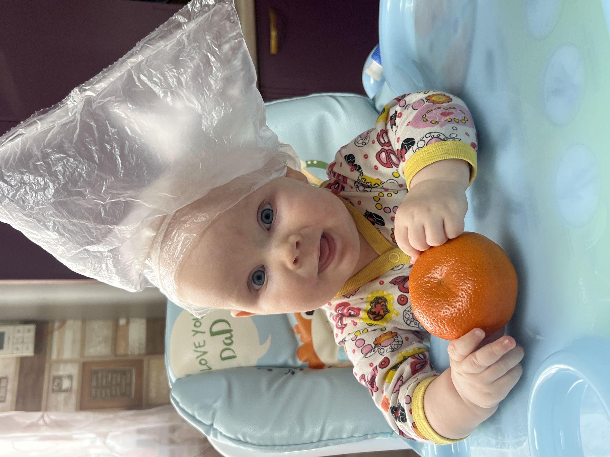An overhead, slightly angled shot captures an infant in a light blue high chair, looking directly at the camera with wide blue eyes and a slight smile, tongue visible. The baby, appearing to be bald, has a crinkled, translucent plastic bag playfully placed on its head, extending over its left shoulder.
The infant is wearing a long-sleeved onesie with a colorful, busy pattern featuring shapes, circles, and what appear to be cartoon characters or objects, with yellow cuffs and collar. Its small hands grasp a vibrant, unpeeled orange. The orange is positioned in the lower right portion of the frame, resting on the light blue surface of the high chair tray, which has subtle circular indentations.
Behind the baby's head, an insert or cushion for the high chair is visible, also in a light blue color, featuring the text "Love You Dad" in green lettering within a speech bubble design. The background beyond the high chair is blurred, showing hints of household interiors, possibly a wall with framed pictures or patterned wallpaper on the left, and a darker, perhaps purple, wall or furniture on the right.
The overall impression is one of a cheerful and curious baby engaged in play, possibly during a mealtime or snack, in an indoor setting, likely a home in Votkinsk, Russia. The time of day is not discernible, but the lighting suggests an indoor environment.