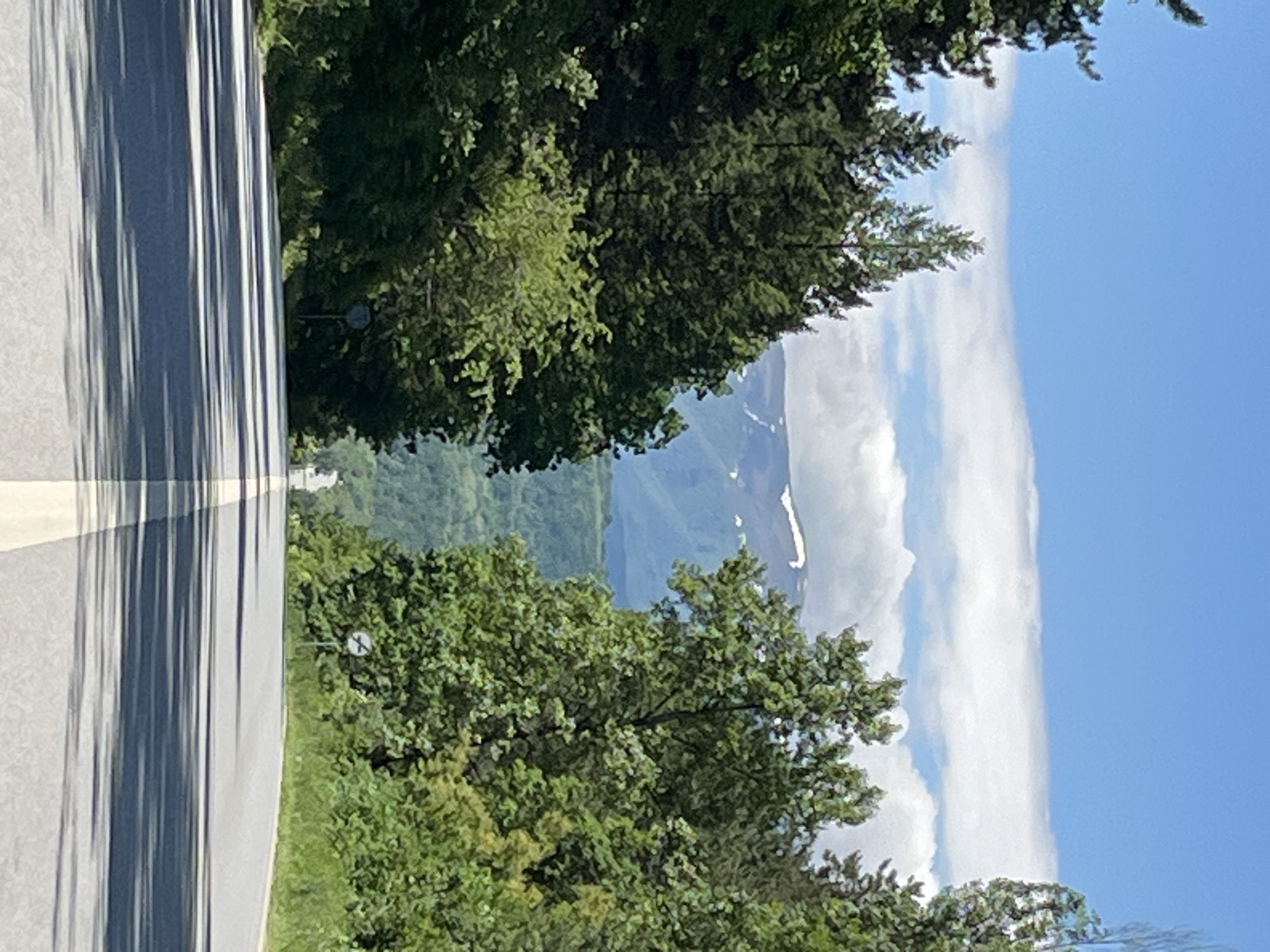The image shows a road leading towards a mountain. The road is paved and has lines painted on it. There are trees on either side of the road. The sky is blue with some clouds. The location is Borovtsi, Bulgaria.