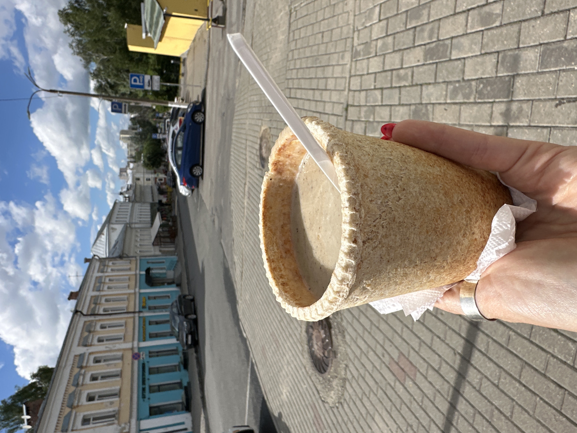 A hand holds a cup made of edible material, filled with a creamy beverage and a white plastic straw. The cup has a textured, grainy surface. The background shows a paved sidewalk with a brick-like pattern, indicating an outdoor urban setting. In the middle ground, a blue car is parked on a street, and beyond that, several multi-story buildings with light-colored facades and blue accents line the street. The sky above is partly cloudy with fluffy white clouds against a blue sky, suggesting a bright, possibly sunny day. A lamppost with cables and a parking sign are also visible. The overall scene appears to be a casual moment outdoors, possibly in a city in Russia given the location context. There are no visible emotions conveyed by the hand.