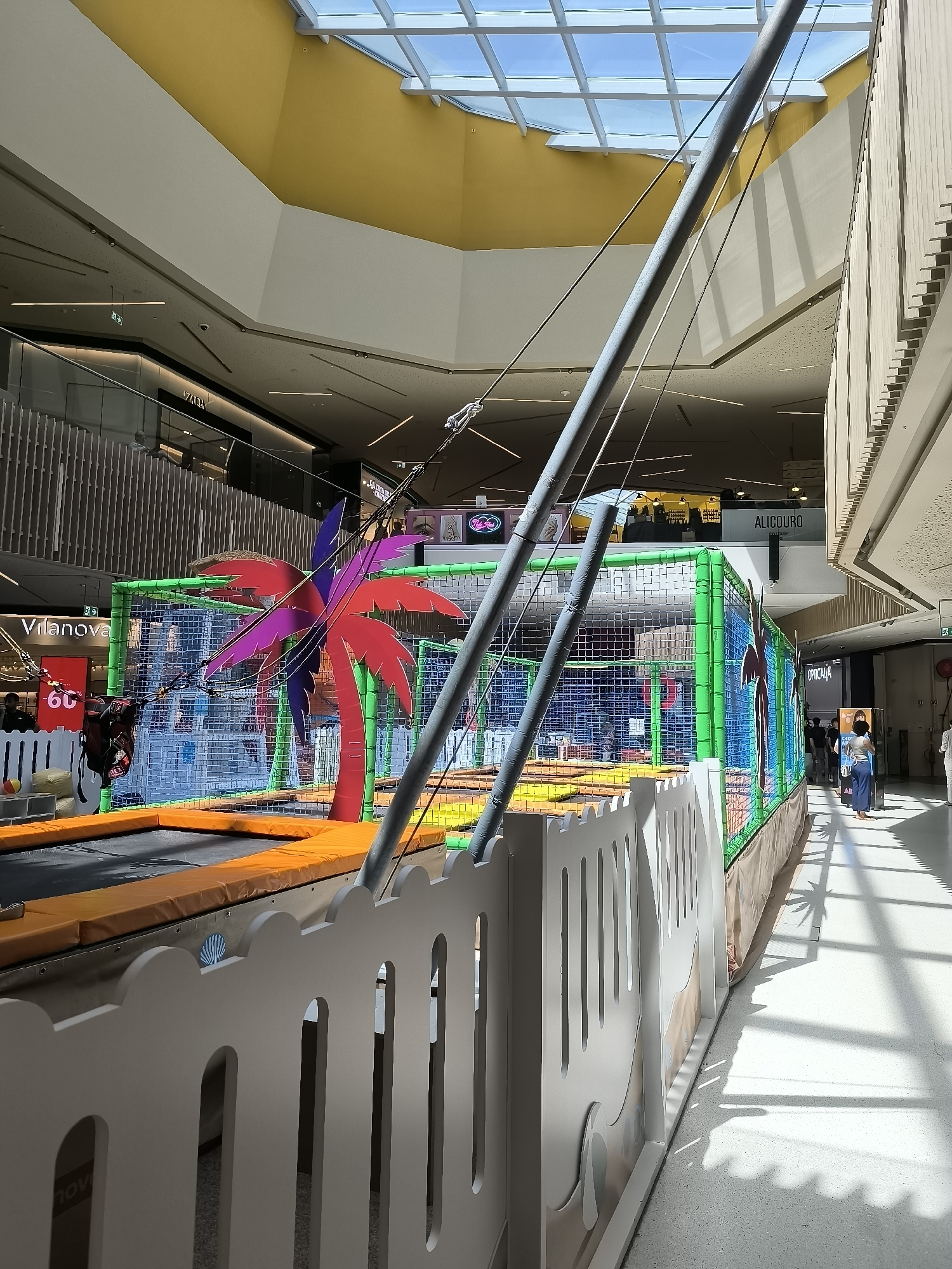 The image shows an indoor children's play area, likely in a shopping mall. The play structure is a trampoline with a safety net enclosure, decorated with a colorful palm tree motif. The environment is bright and airy, with a skylight overhead that casts distinct shadows on the floor. In the background, there are shop fronts and other shoppers walking by. The specific location is the Parque Nascente shopping center in Setúbal, Portugal. A white, decorative fence with cutouts of cartoon animals encloses the play area. Several people, including children, are present, some actively using the play equipment and others observing. The lighting suggests it is daytime.