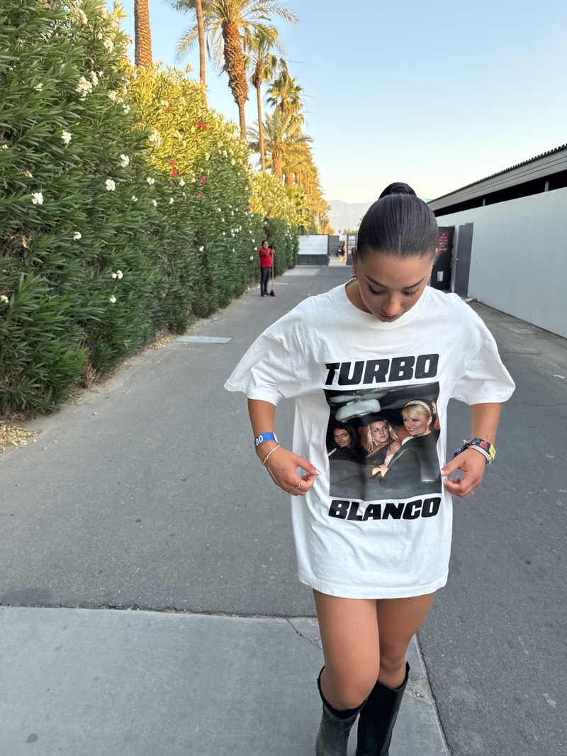 A young woman stands on a sidewalk in Indio, California. She is wearing a large white t-shirt with a picture of "Turbo Blanco" on it and tall black boots. Her hair is pulled back in a high ponytail.
Behind her, there's a row of tall green bushes with white flowers, lined up along the sidewalk. Beyond that, there are palm trees reaching into the sky and some low-lying mountains in the distance. In the background, a person is sweeping. The sky is a clear, light blue.