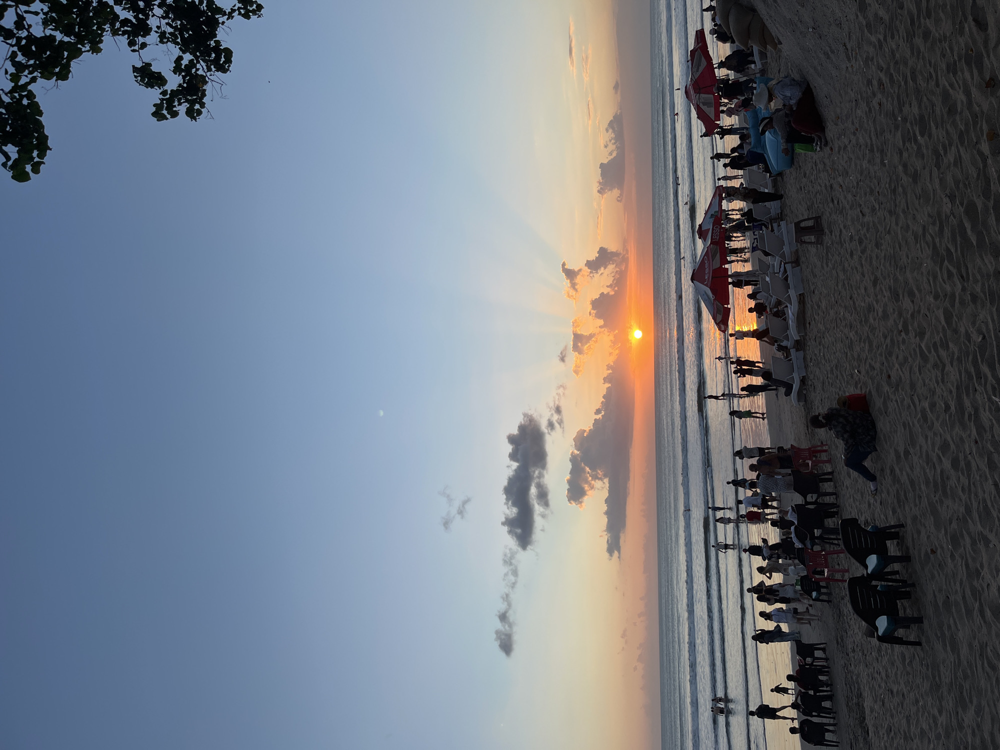 The image depicts a crowded beach scene in Kuta, Indonesia, during sunset. The sun, a bright orange orb, is low on the horizon, casting golden rays that fan out into the pale blue sky. Several clouds, illuminated by the setting sun, are scattered across the sky, with some appearing as wispy trails and others as more substantial formations. The ocean features gentle waves rolling towards the shore, with a few surfers visible in the water.
On the sandy beach, numerous people are gathered. Some are wading in the shallow water, while others are standing and walking along the shoreline. Further back from the water's edge, beach chairs and umbrellas are set up, indicating a recreational area. Groups of people are seen sitting and conversing on the sand, some in chairs and others on beach lounges. Beach umbrellas, predominantly red with white logos, provide shade for some of the seated individuals. The sand is textured, with footprints and ripples visible. In the foreground, several empty plastic chairs are arranged on the sand, suggesting a seating area that may have recently been occupied or is waiting for patrons. The overall atmosphere appears to be one of relaxation and enjoyment of the evening scenery. The foliage of a tree is visible in the upper right corner of the frame, silhouetted against the sky.