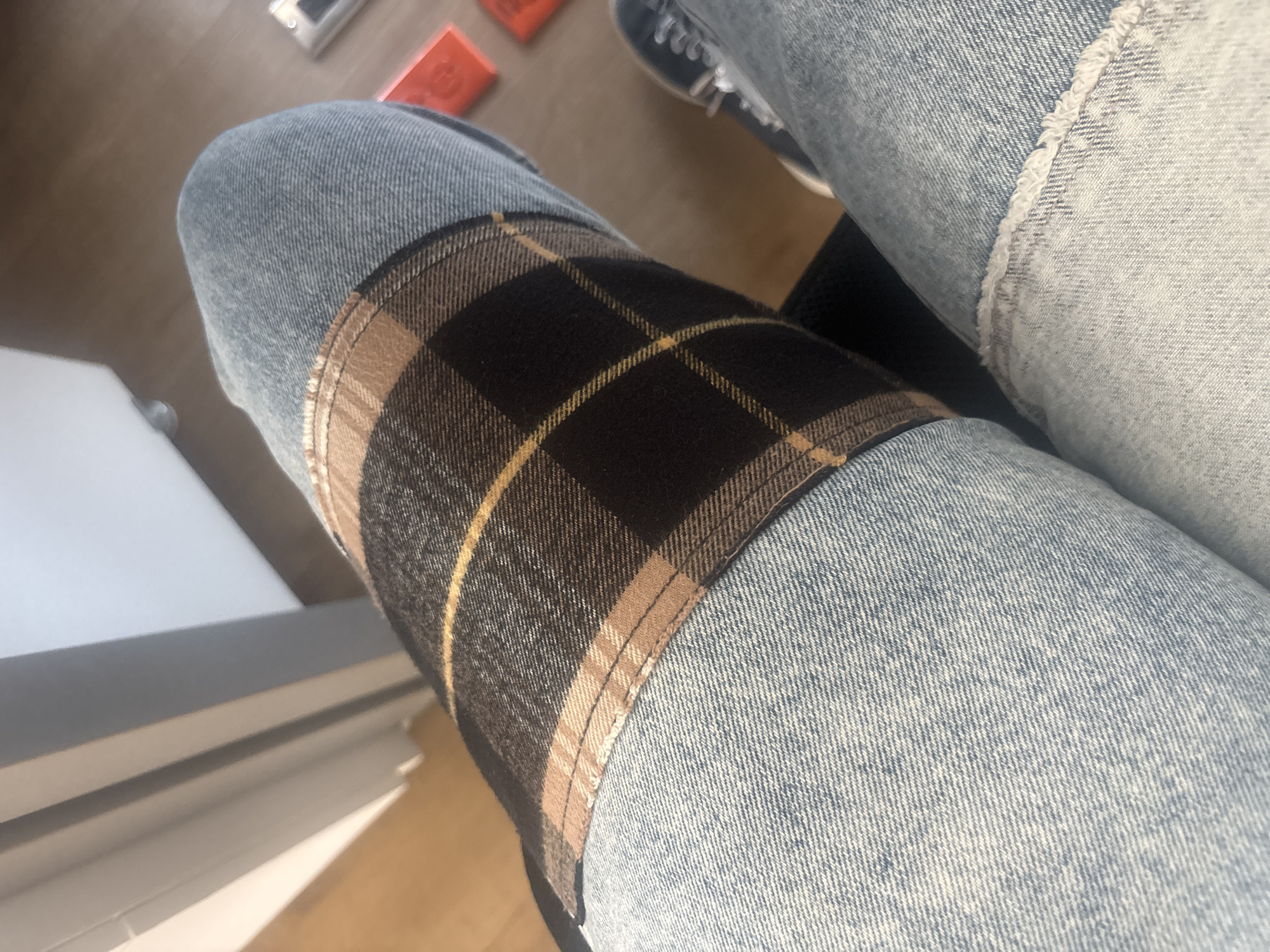 This close-up image, set in Buenavista, Mexico, features a person's legs clad in light-wash denim jeans, seen from a high-angle perspective. The individual appears to be seated or resting their legs in an indoor environment. A notable detail on the jeans is a large, rectangular plaid fabric panel sewn onto the knee of the left leg (from the viewer's perspective). This plaid pattern is composed of dark, possibly black or deep brown, interwoven with intersecting stripes of light brown/beige and thin golden-yellow lines. The stitching around this patch is clearly visible.
In the background, a warm-toned wooden floor is visible, sparsely adorned with a few small, light-colored rectangular objects and some red items. The presence of strong highlights and shadows indicates bright, natural daylight, suggesting it is a sunny day. On the right side of the frame, the front portion of a shoe, likely a sneaker with white laces, is partially visible. To the left, a segment of a white or light-colored furniture piece can be seen. No specific activity is occurring, and there is no visible text or discernible emotions.
