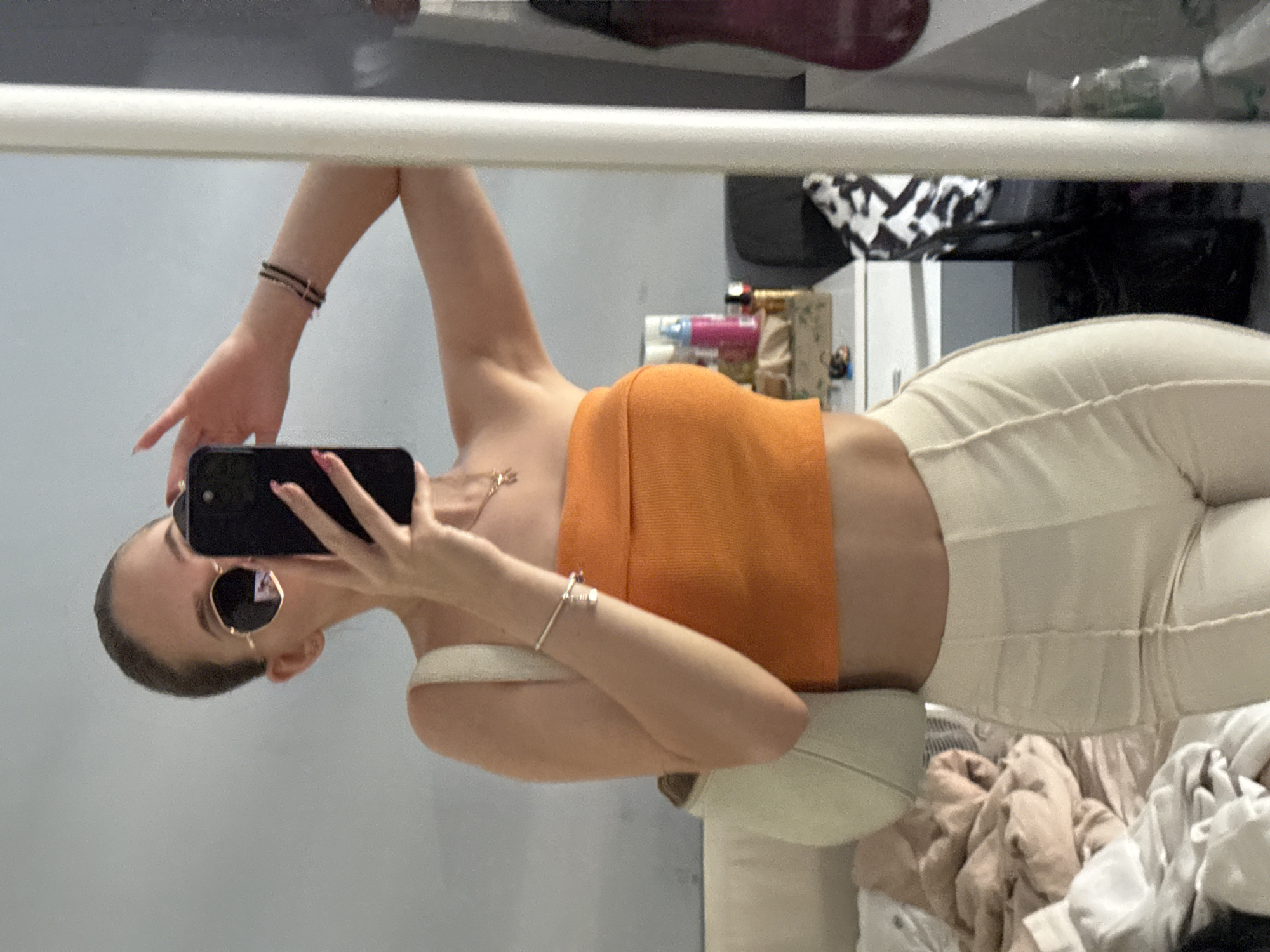 A young woman is taking a mirror selfie in what appears to be a bedroom. She is wearing an orange strapless top and cream-colored pants. She has on round sunglasses and is holding a smartphone in her right hand to take the picture. A white handbag is slung over her left shoulder. The background shows a bed with rumpled bedding and a dresser with various items on top, including bottles and a tissue box. A full-length mirror is prominently featured, reflecting the scene. The overall impression is a casual, personal moment captured at home. There is no indication this scene is located in Sofia, Bulgaria.