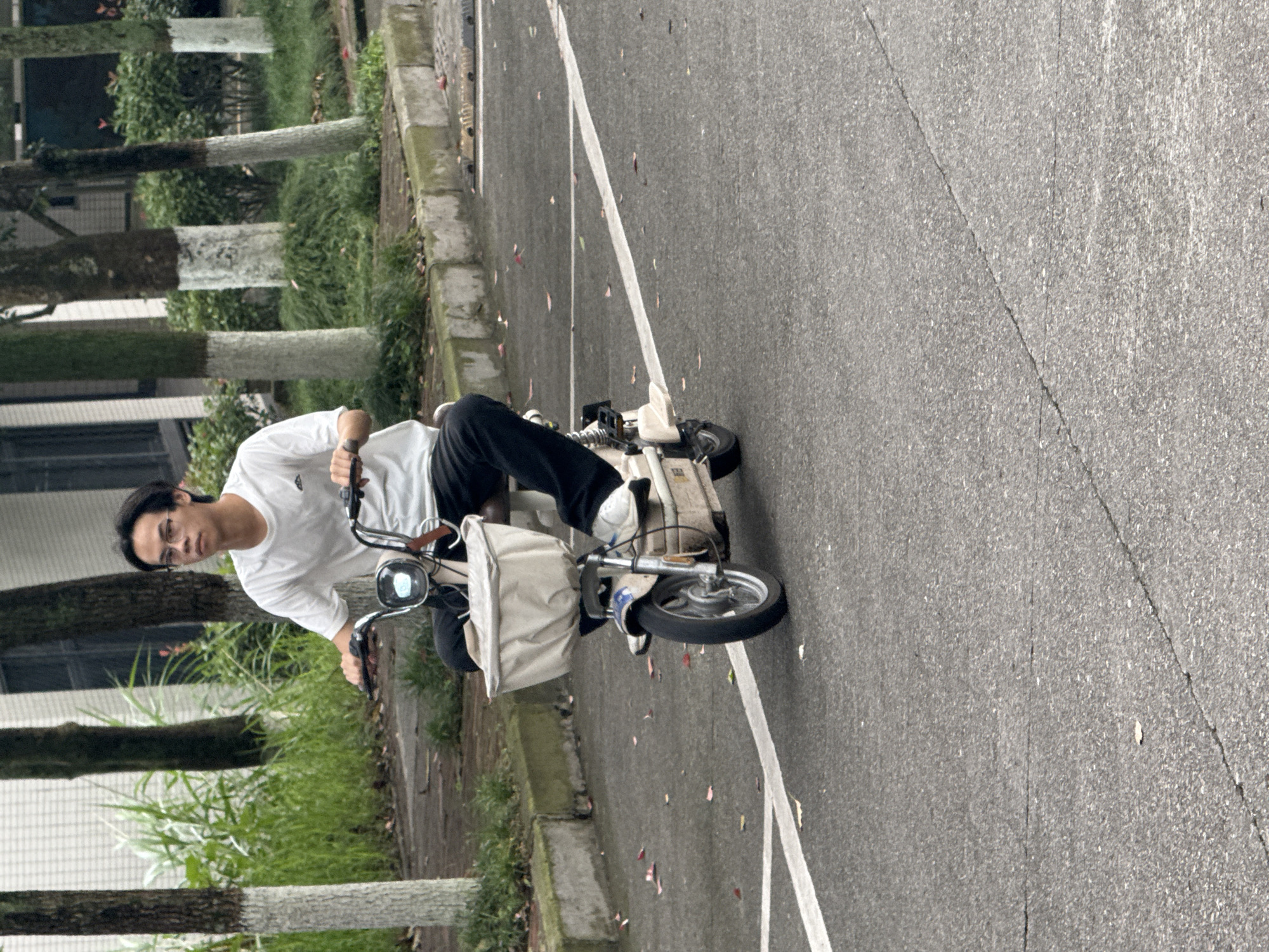 The image shows a person riding a small, possibly electric, scooter on an asphalt road with white lane markings. The person is wearing a white t-shirt, dark pants, and glasses. The scooter has a basket attached to the front. To the left of the road, there is a curb and a grassy area with trees. The setting seems to be a residential area, likely in a suburban or urban environment. There is no specific detail that can identify the specific location in Jiangjin, China.
