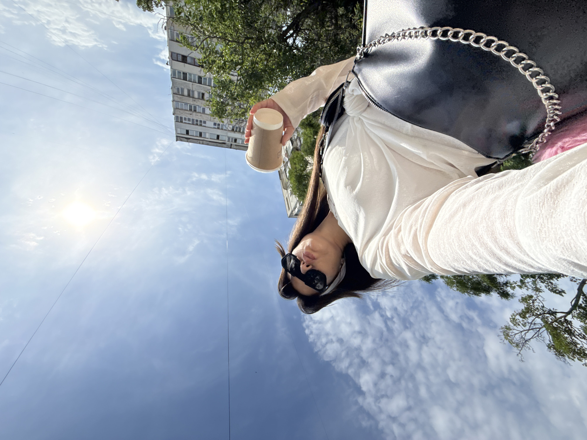 A low-angle, wide shot captures a woman holding a coffee cup against a bright, sunny sky with scattered clouds. She is wearing sunglasses, a white long-sleeved shirt, and a black purse with a silver chain strap. Behind her, a tall, multi-story building with many windows is visible, along with trees. Power lines are stretched across the sky. The woman appears to be taking a selfie, with her face angled upwards. The environment suggests an urban setting, likely outdoors during the daytime.