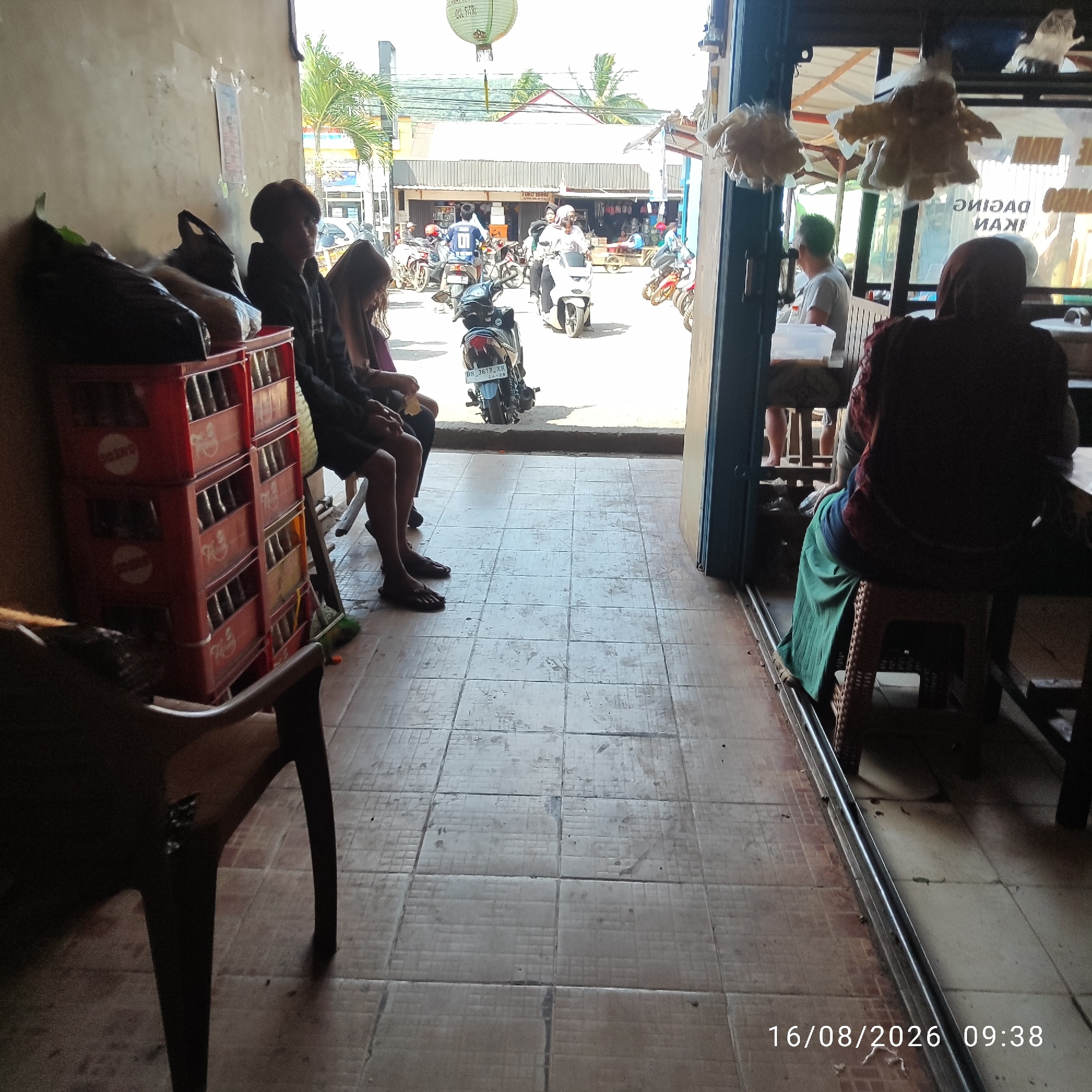 In the city of Pembaharuan, Indonesia, a street-level view captures a moment outside a small establishment. In the foreground, a person sits on a wooden chair in shadow, facing away from the camera, with their legs crossed. To their right, another person, also seated, is partially visible, wearing a dark headscarf and a long green skirt. They are seated on a woven plastic stool.
The interior of the establishment is dimly lit, with shelves holding stacked red crates, possibly for beverages, visible on the left. Further inside, a man with short dark hair is seated, looking to his left, while a woman with long dark hair sits beside him.
The exterior scene is bustling with activity. A street is visible with several motorbikes parked and some in motion. People are seen riding motorbikes, including a person in a white top and headscarf on a white motorbike, and a person in a blue shirt with the number '0' on their back on a dark motorbike. In the background, buildings with corrugated metal roofs line the street, suggesting a commercial area. A white paper lantern hangs from a wire. The ground is tiled, showing signs of wear and tear. A timestamp in the lower right corner indicates the image was taken on 16/08/2026 at 09:38.
