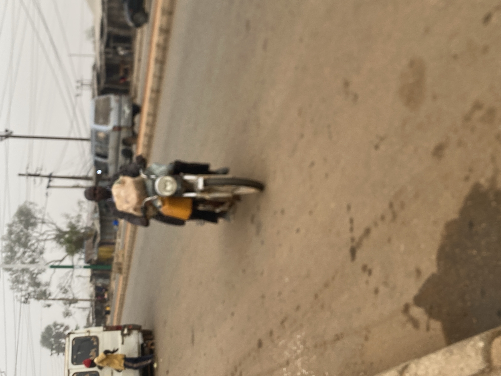 The image depicts a street scene in what appears to be Bukuru, Nigeria. A motorcycle with two riders is traveling on the paved road. The motorcycle has a large, light-colored bag strapped to the front. Other vehicles, including vans and what looks like a small bus, are parked or moving alongside the road. Power lines are visible above, adding to the urban character of the location. A few people are visible near a white van. The overall environment seems overcast or hazy.