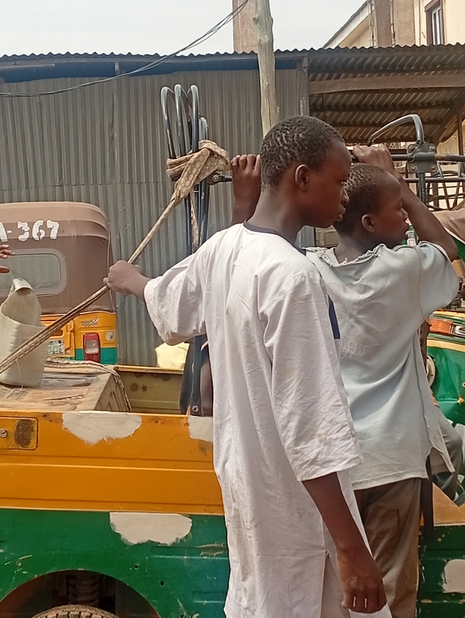 Two boys stand alongside a green and yellow vehicle in Jos, Nigeria. One boy, facing away, wears a white shirt and holds a long wooden stick, while the other wears a pale blue shirt and is partially obscured. The vehicle appears to be undergoing repair, with patches of white filler visible on the body. Behind them is another similar vehicle, a corrugated metal wall, and parts of buildings. Metal bars are bundled on the vehicle.