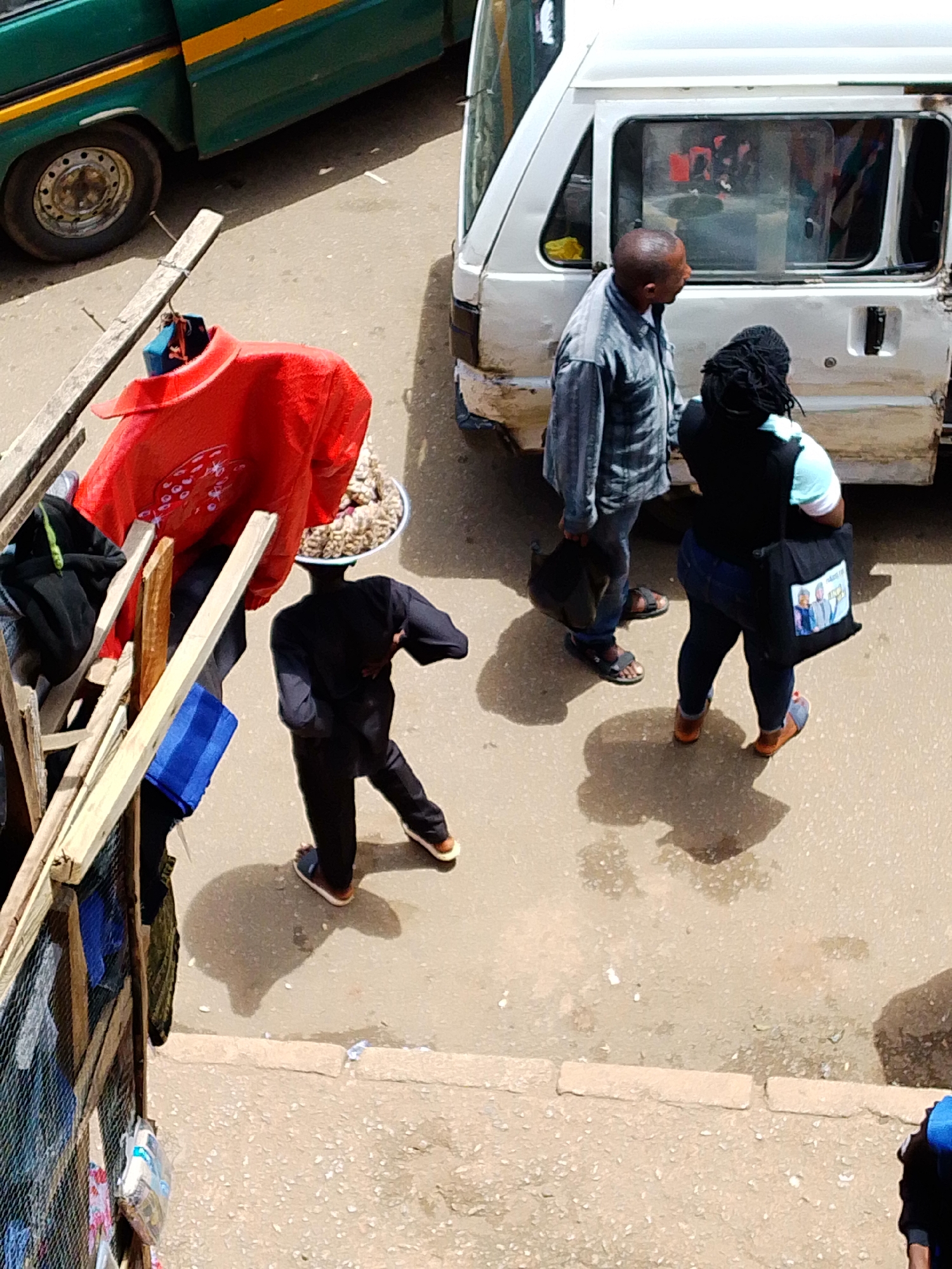 A medium-angle aerial shot shows a busy street scene with two people standing near a white van and a green bus. A person in a black outfit with a bowl of goods on their head walks away from the viewer. Another person, dressed in a light-colored button-down shirt and dark pants, stands facing a person in a dark vest and light blue shirt. The white van is open at the back. A red jacket is draped over a wooden structure on the left side of the image. The ground is asphalt, and shadows of the people and objects are visible.