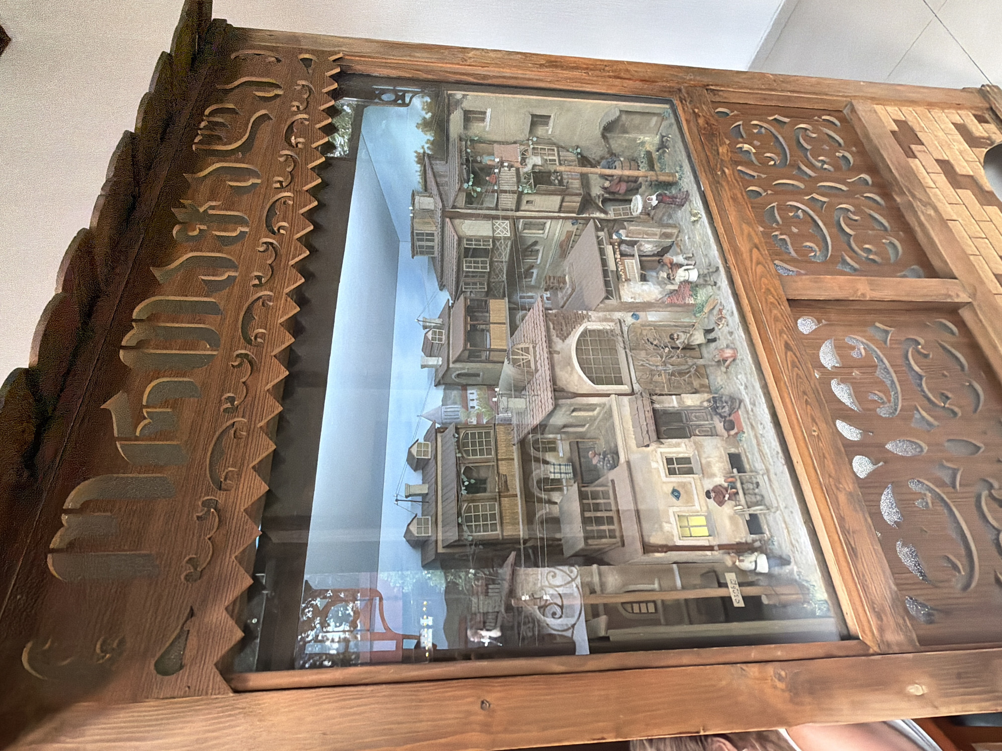 The image shows a diorama depicting a street scene in the city of Tbilisi, Georgia. The diorama is housed within a wooden display case with carved decorative elements. The street scene itself features several multi-story buildings with architectural details characteristic of old Tbilisi, including wooden balconies and ornate windows. Miniature figures are positioned throughout the scene, engaged in various activities such as standing, sitting, and interacting with their surroundings. The diorama includes elements like street signs, barrels, and even a small dog, contributing to a realistic portrayal of urban life. The background of the diorama depicts a painted sky and distant hills. The overall impression is a detailed miniature representation of a historical urban environment.