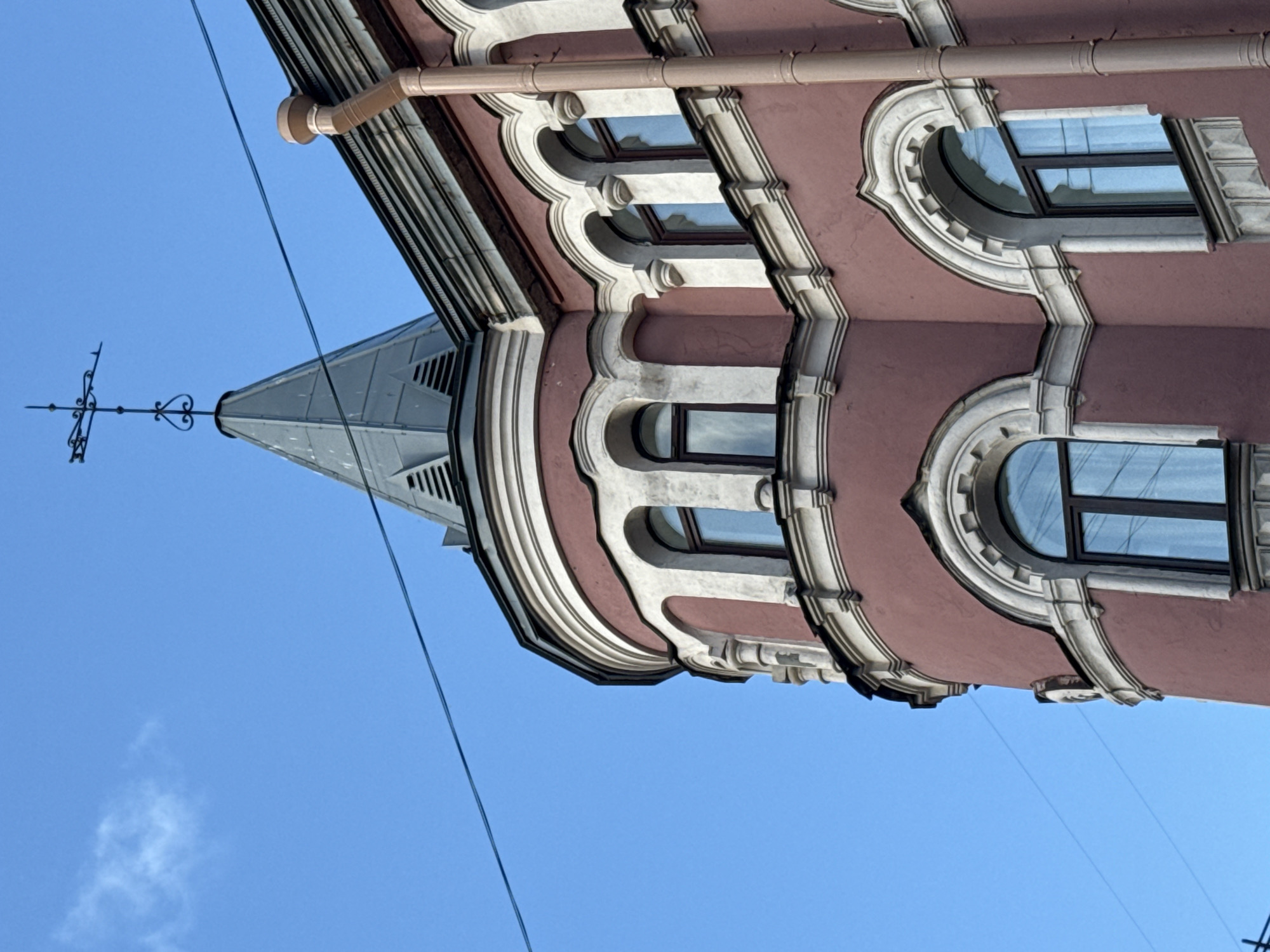 The image shows a close-up, low-angle view of the upper section of a historic building in Saint Petersburg, Russia, against a clear blue sky. The building features a prominent rounded tower with arched windows, adorned with decorative white trim. The facade is painted a muted reddish-pink color, with bands of white plasterwork and architectural details.
Atop the tower is a gray, conical roof topped with a wrought-iron weather vane shaped like a cross with decorative elements. A light brown downspout runs vertically along the side of the building, disappearing below the frame. In the background, thin black wires stretch across the sky, suggesting the presence of overhead power or telephone lines. The architecture suggests a late 19th or early 20th-century style common in Saint Petersburg. The image is a still photograph with no discernible activity or people present.