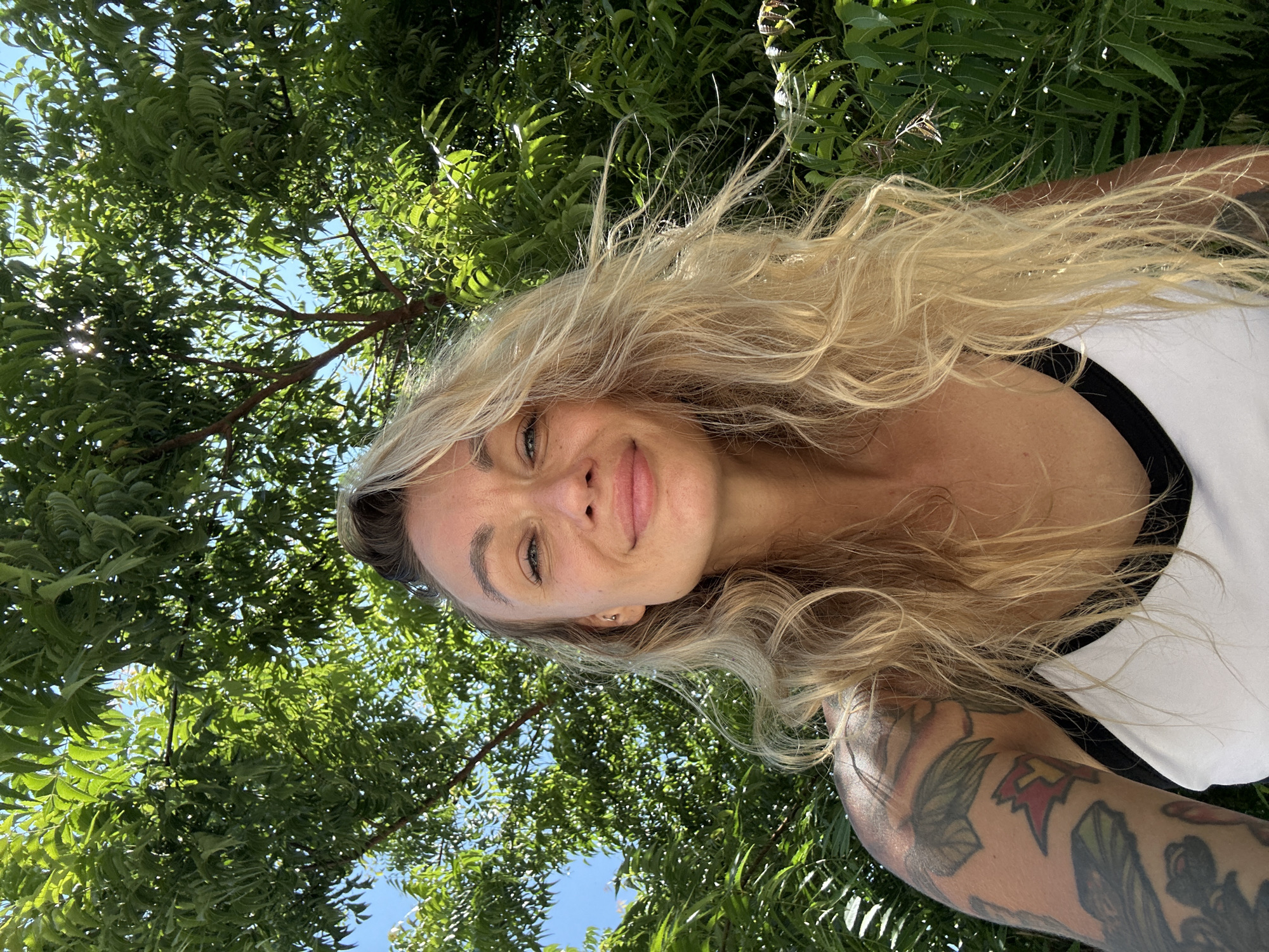 A medium shot captured from a low angle shows a woman with blonde, wavy hair and a subtle smile, looking directly at the camera. She is wearing a white tank top with a black trim. Her left arm, visible on the left side of the frame, is extensively tattooed. The background is dominated by lush green foliage, suggesting an outdoor setting. The foliage appears to be from a tree with dense leaves, and patches of blue sky are visible through the canopy. The lighting indicates it is daytime, with sunlight filtering through the leaves, creating dappled shadows. The image does not provide any specific visual cues to pinpoint its location within the city of Mũi Né, Vietnam; it depicts a common natural environment.
