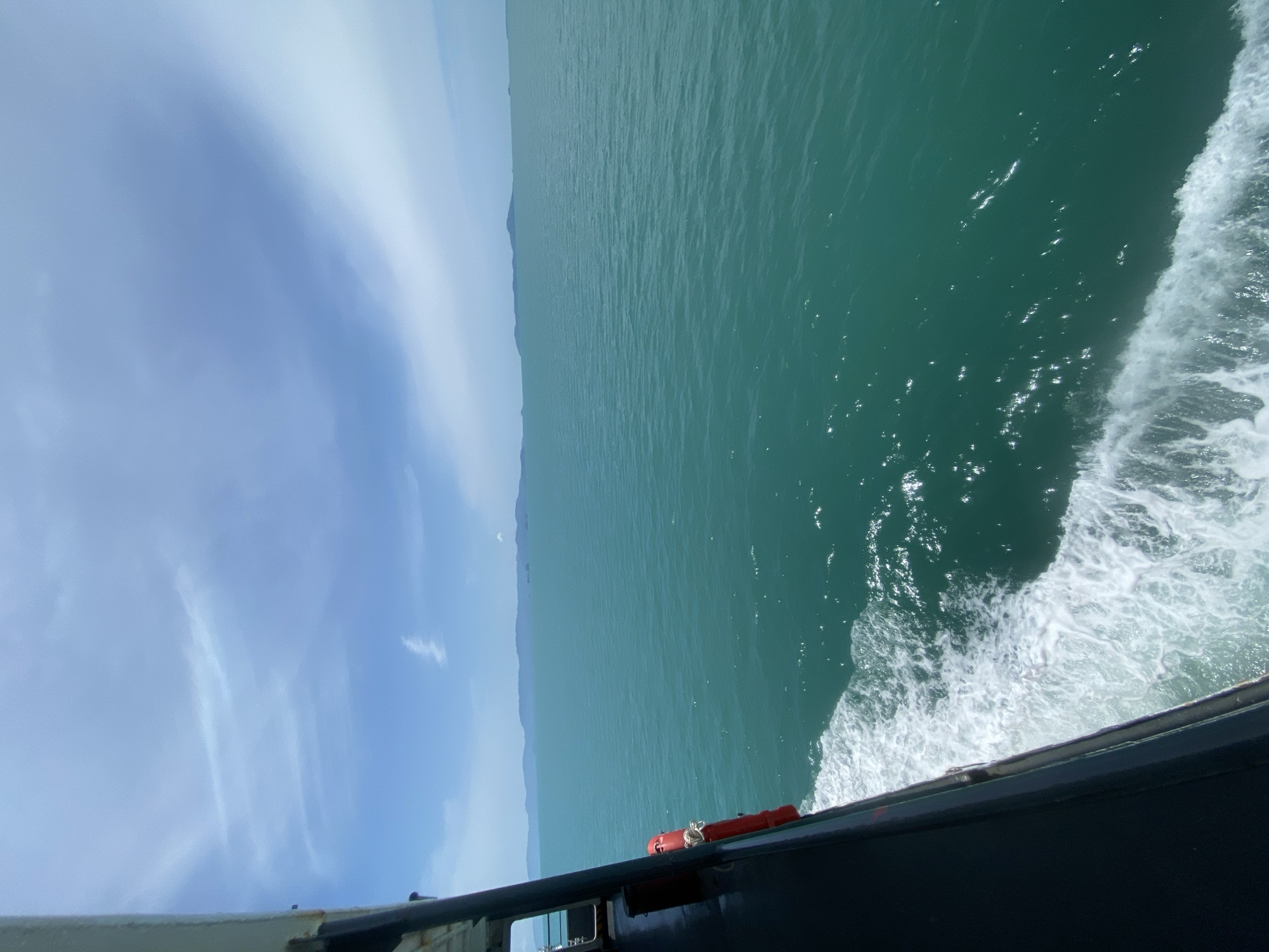 The image depicts a view from the side of a vessel navigating through calm, turquoise water. The vessel, which appears to be a ferry or a similar type of boat, is partially visible on the left side of the frame, with a dark hull and a railing. A bright orange life preserver is attached to the side of the boat. The water is a vibrant teal color and shows gentle ripples and reflections of sunlight. A white wake trails behind the vessel, indicating movement. In the distance, a chain of islands or the mainland coastline is visible as a low, undulating silhouette against the horizon. The sky above is a pale blue with scattered, wispy clouds. The scene suggests a journey on the sea, likely in a tropical or subtropical region. The location is specified as Thong Nian, Thailand, which is known for its coastal scenery and maritime activities.