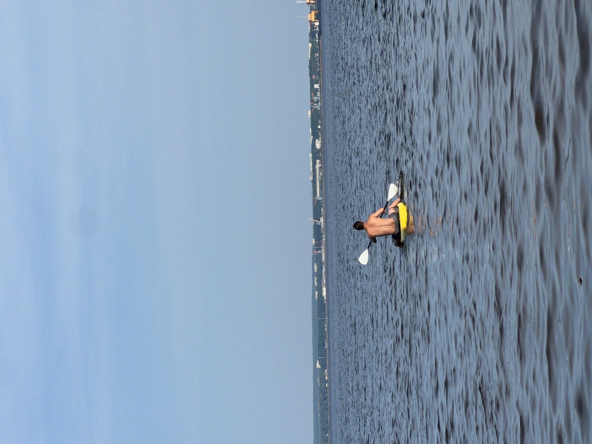 A person is paddleboarding on a large body of water. The person is facing away from the camera, wearing black shorts. They are on a yellow paddleboard with a white paddle. In the background, there is a city skyline with cranes and buildings. The water is dark blue and choppy. The sky is a clear, pale blue. The image appears to have been taken on a bright, sunny day. This scene is located in Noviy, Russia.