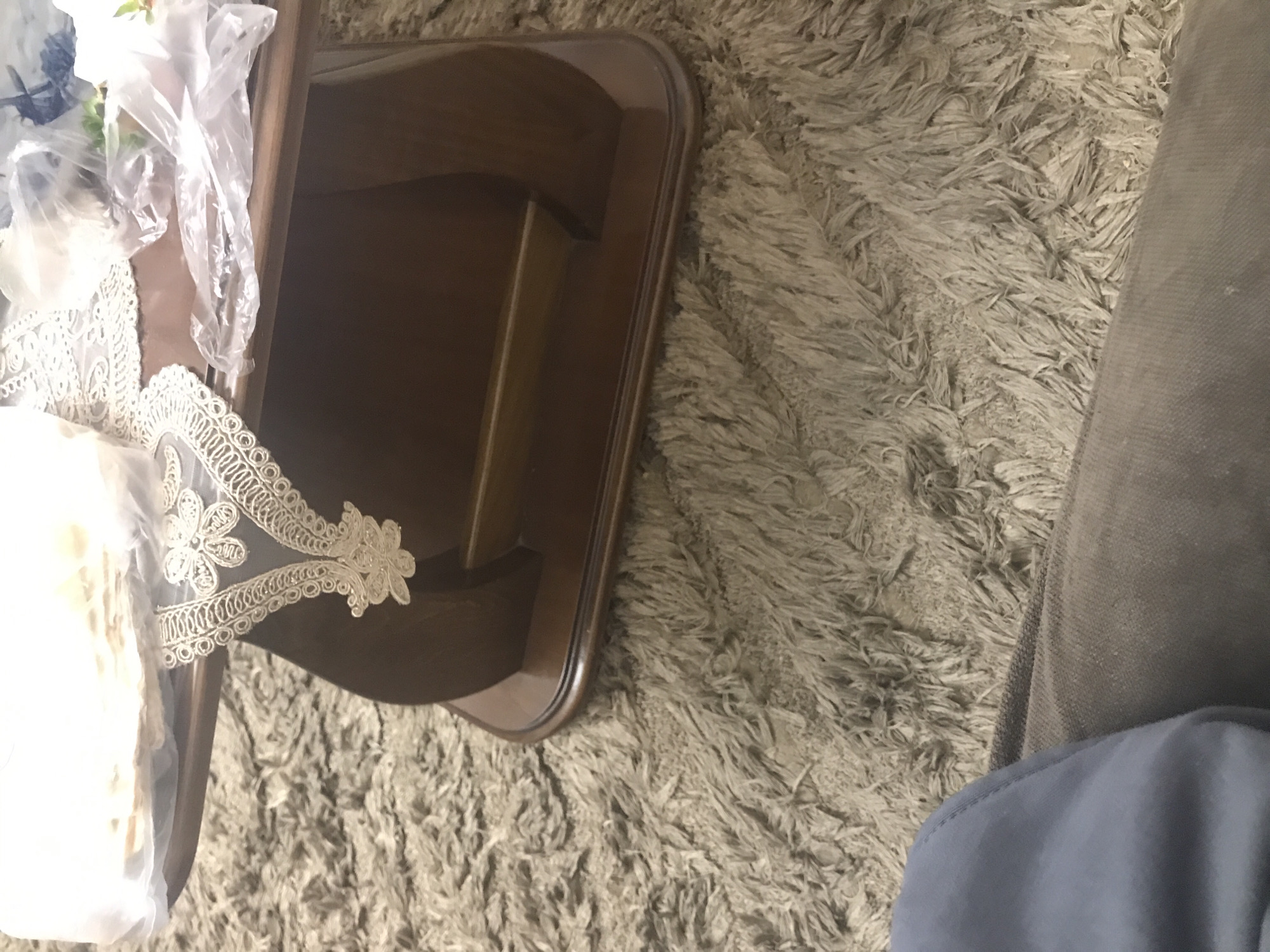 The image shows a close-up, overhead view of a brown, two-tiered table partially covered with lace and plastic-wrapped items. The table rests on a shaggy, light-colored rug. The edge of a gray fabric material is visible on the right side of the image. There is no information that supports that the image location is in Yerevan, Armenia.