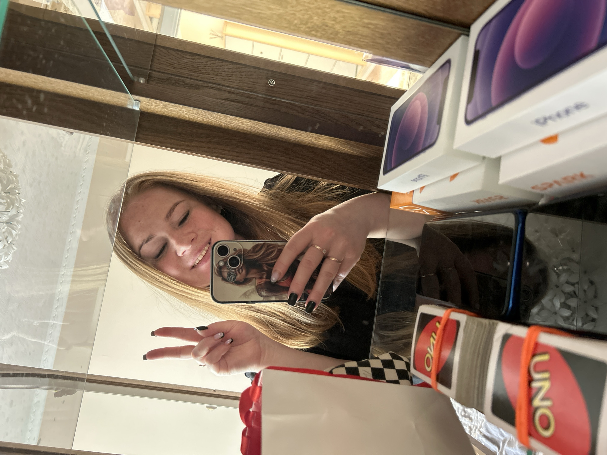 A young woman with long blonde hair takes a selfie in a mirror, likely in a store located in the city of Chaikovsky, Russia, based on the presence of Russian-language "SPARK" branding on the boxes. She holds a phone with a decorative case, making a peace sign with her left hand. The reflection shows shelves stocked with iPhone boxes and packs of UNO cards, some secured with orange rubber bands. A shopping bag with red accents is visible in the foreground. The scene is brightly lit.