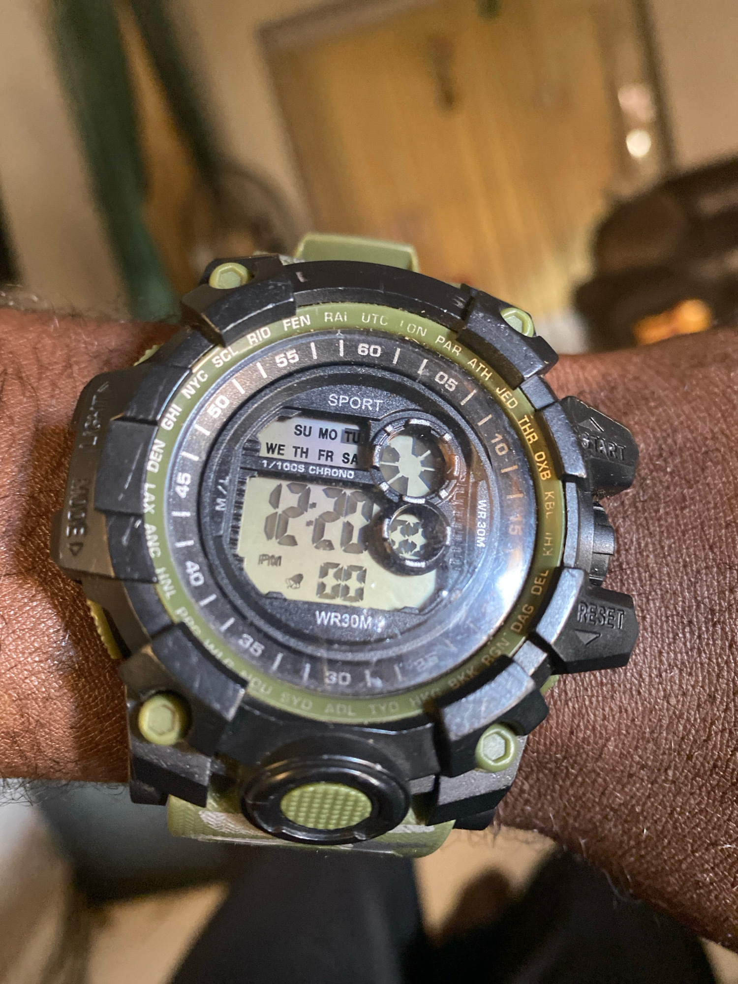 The image shows a close-up of a digital watch on a person's wrist. The watch is black and green with a digital display. The time displayed is 12:22. The watch face has various markings, including city abbreviations and numbers. The background is blurry and appears to be indoors. The wrist is covered in skin, with visible pores and hair. The image quality is adequate for identifying the watch and its display. There is no indication of the location in the city of Jos, Nigeria.