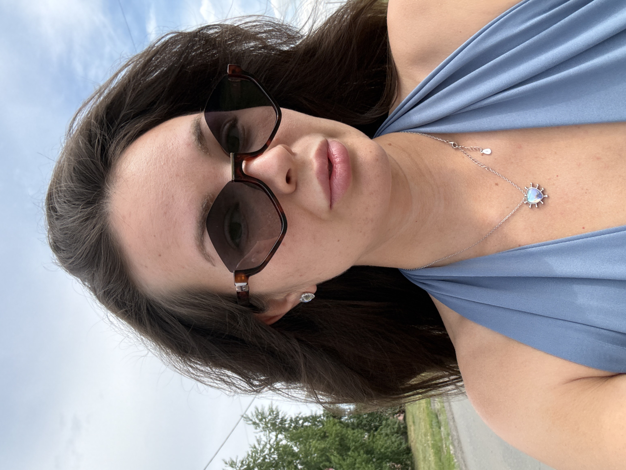 A woman with long, dark brown hair and prominent cheekbones is pictured in a close-up, outdoor shot. She is wearing large, angular sunglasses with a tortoise-shell pattern and a blue halter top. A silver necklace with a heart-shaped pendant adorned with what appears to be a milky, iridescent stone is visible around her neck. She is also wearing small, stud earrings. The background suggests a semi-rural setting with green trees and a cloudy sky. The location is specified as Bely Yar, Russia.
