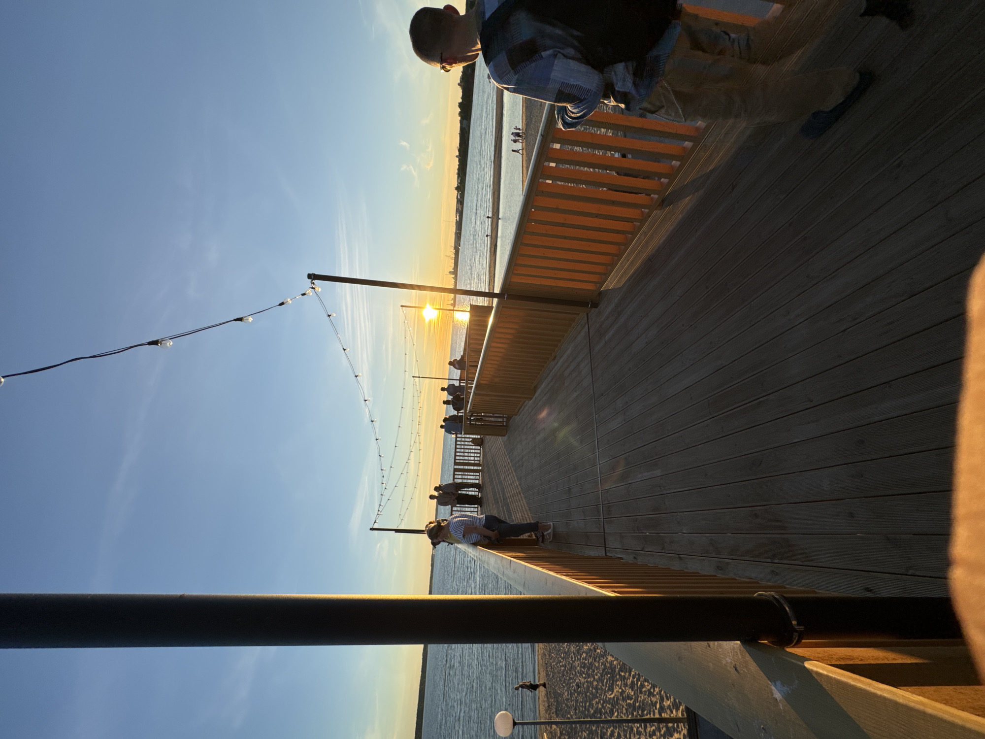 The image shows a wooden pier on a beach at sunset. The sky is a clear blue with a few wispy clouds. The sun is setting over the water, casting a warm glow on the scene. A group of people are standing on the pier, looking out at the sunset. A man with a backpack is standing on the right side of the pier, looking away from the camera. On the beach, a person is walking by the water, and further down the beach, a group of people are gathered. The pier has wooden planks and a wooden railing with orange vertical slats. String lights are hung above the pier. The environment appears to be a public recreational area.