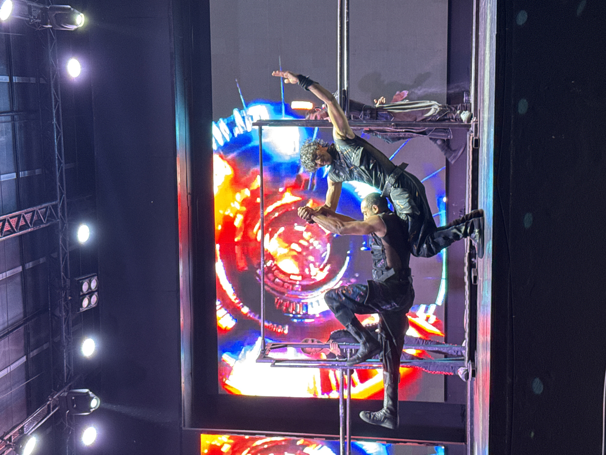 In the city of Manavgat, Turkey, two male acrobats perform on a stage in front of a large LED screen. The acrobat on the left is upside down, supported by the acrobat on the right, who is balancing on one arm. Both performers are wearing dark, fitted athletic wear. The LED screen behind them displays a vibrant, abstract pattern of red, yellow, blue, and white. The stage is illuminated by stage lighting. A third person, also male, dressed in darker clothing, stands behind the acrobats, holding onto the metal bars that the acrobats are using for their performance.