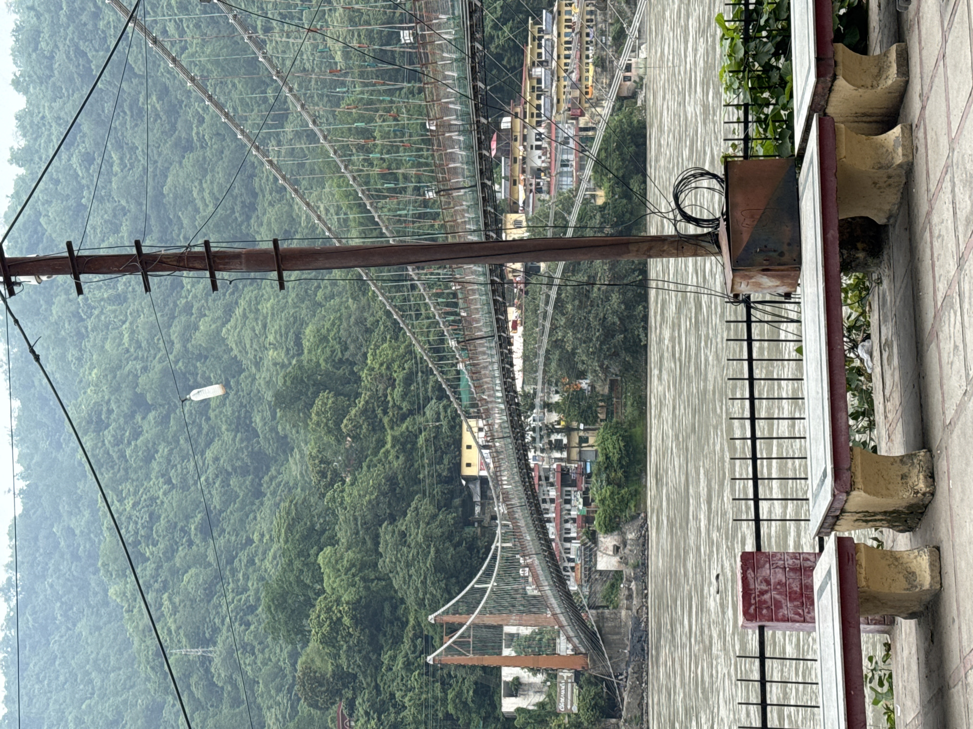 The image depicts the Lakshman Jhula suspension bridge in Rishikesh, India, spanning the Ganges River. In the foreground, two stone benches with red and yellow accents sit on a tiled pathway, separated by a red brick pillar. Behind the benches, a black metal fence lines the riverbank, beyond which the muddy, turbulent waters of the Ganges flow. A rusty utility pole with coiled wires stands to the right of the benches, obscuring part of the bridge. The bridge itself is a long, metal suspension structure with a pedestrian walkway, its orange-colored support towers visible. On the far bank of the river, buildings with yellow and red roofs ascend the densely forested hillside, characteristic of Rishikesh. The sky is overcast, and the overall atmosphere is muted, with lush green foliage dominating the background. There are no visible people or animals in the scene.