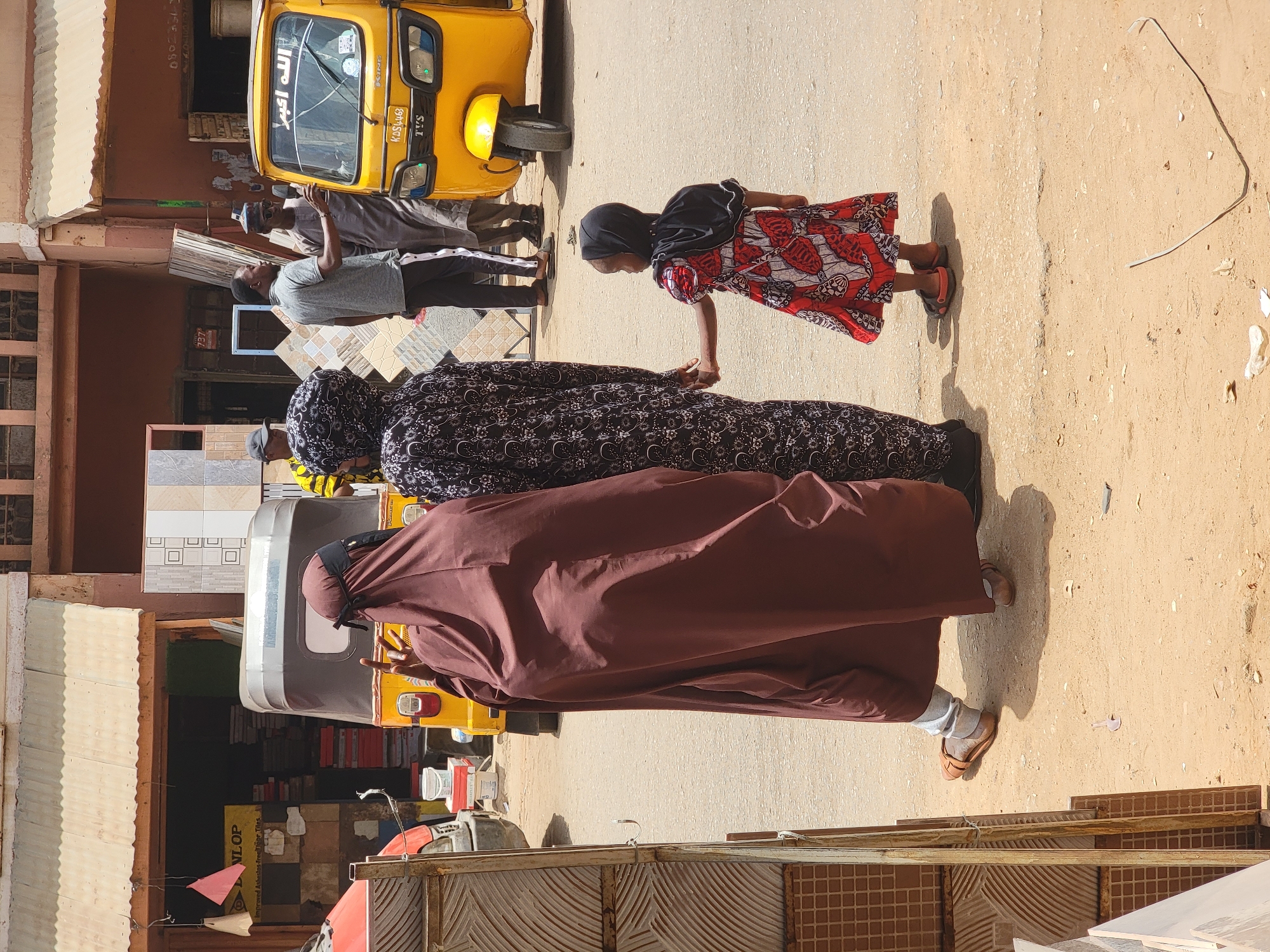The image depicts a street scene in Maiburiji, Nigeria. A group of people, including women and children, are seen walking along a dusty road. One woman is wearing a brown robe while another woman is in a black and white patterned outfit. A child in a red and white patterned dress is holding the hand of one of the women. Two men are near a yellow tuk-tuk, also known as a Keke Napep, a common form of transportation in Nigeria. Commercial establishments, including a shop selling tiles, are visible in the background.
