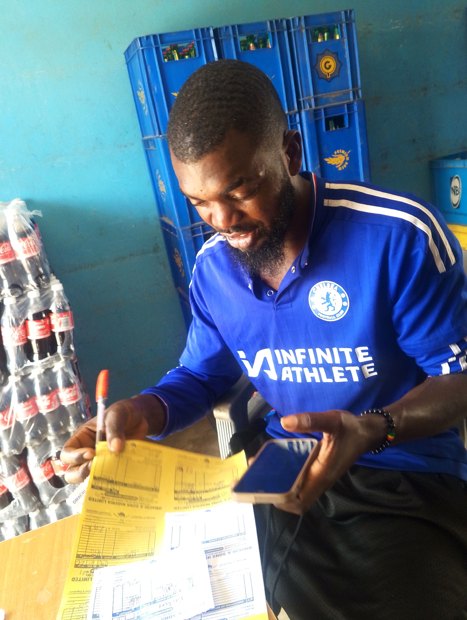 A man with a beard wearing a blue Chelsea football club shirt and black shorts is sitting at a wooden table, looking down at yellow papers and a smartphone. He is holding a pen in his right hand and the phone in his left. In the background, there are stacks of blue plastic crates and several plastic bottles of Coca-Cola. The scene appears to be indoors, with a light blue wall in the background. The image was taken in Jos, Nigeria.