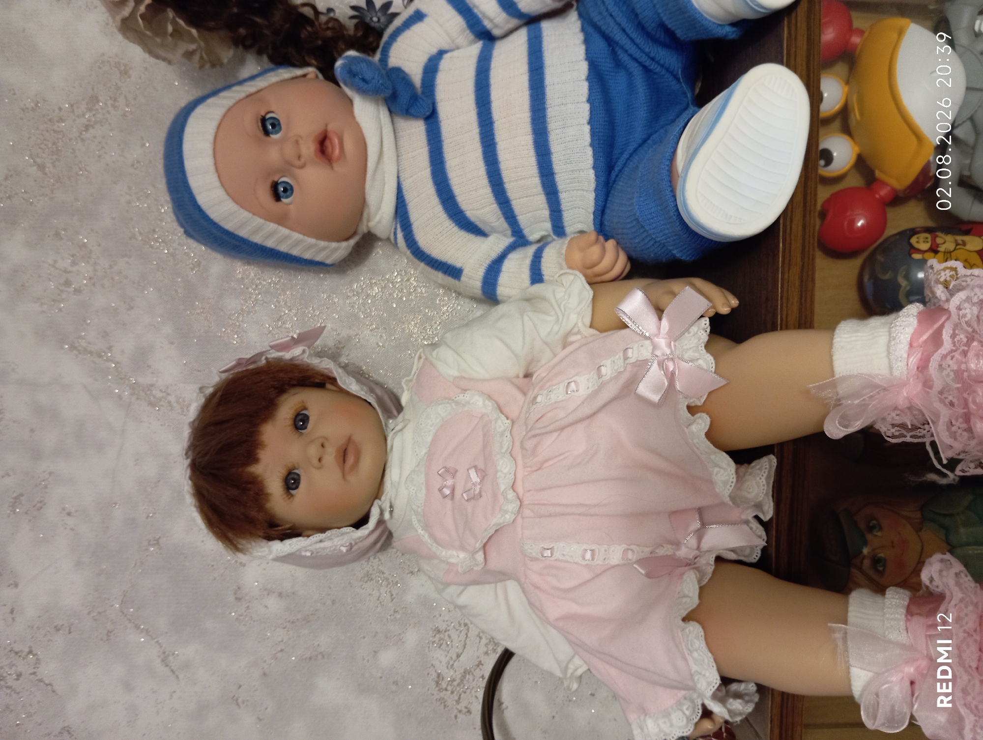 The image displays two dolls seated side-by-side on a shelf. The doll on the left is a baby doll with brown hair, wearing a pink dress with white lace trim and a matching bonnet. The doll on the right is a baby doll wearing a blue and white striped sweater and blue pants, with a blue and white striped hat. The background appears to be a textured wall. Below the dolls, a small wooden doll with blonde hair and a green outfit is visible, along with a red crab toy and other toy items. The timestamp "02.08.2026 20:39" and the text "REDMI 12" are present in the bottom corners of the image. There is no information in the image to suggest a location in Oryol, Russia.