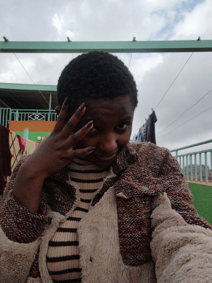 A person with short, dark hair and painted nails poses outdoors. The individual wears a striped top and a textured jacket with a fuzzy lining. The background features a cloudy sky, a green structure overhead, and a balcony railing. Laundry hangs on a line behind the person. The person's hand is near their face, partially obscuring their eye.