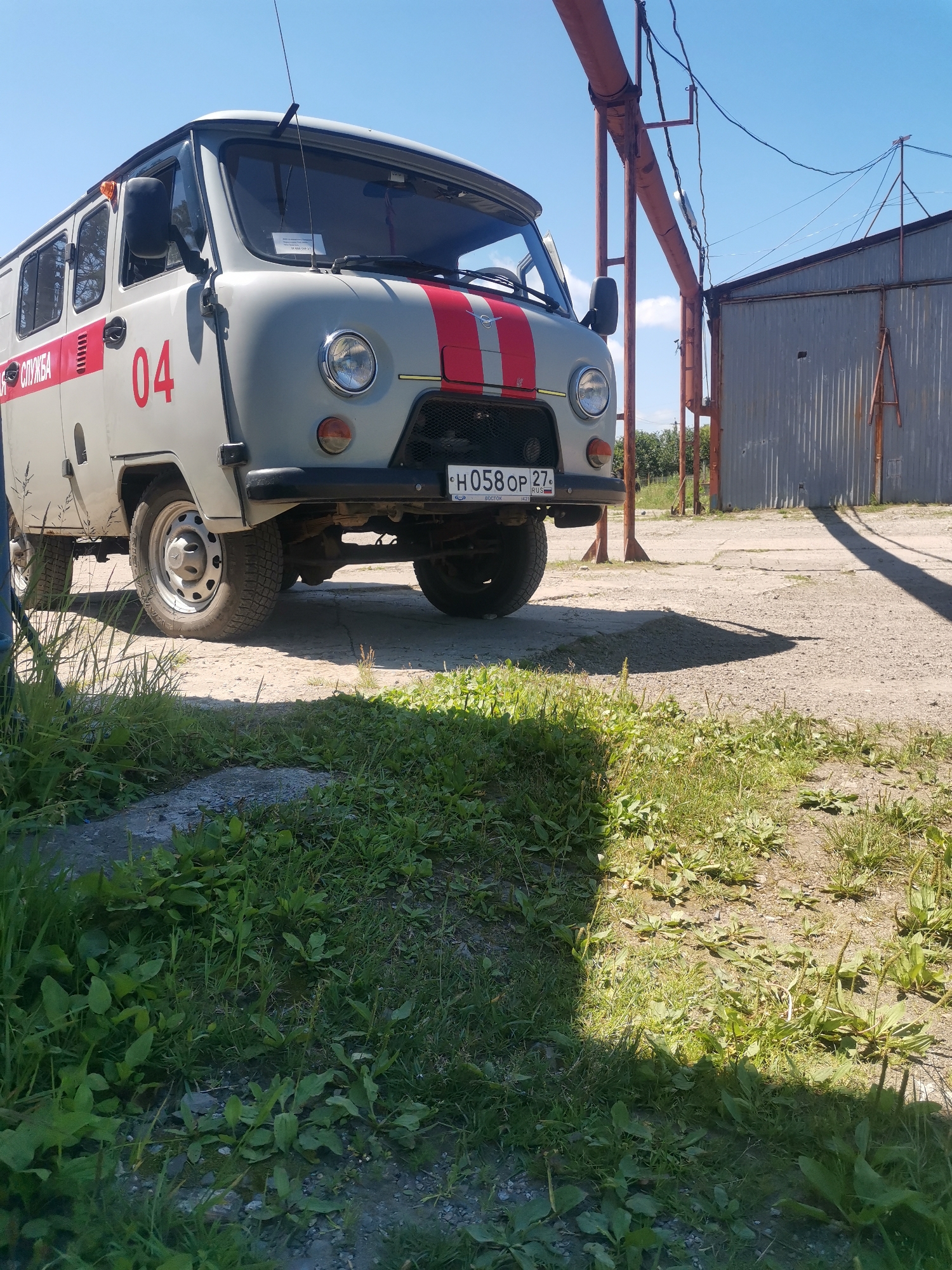 A light gray UAZ-452 van, marked with "СЛУЖБА" (Service) and the number "04" in red, is parked on a gravelly surface near a corrugated metal building. The van has red stripes on the front grille and sides. A license plate, partially visible, reads "H058OP 27 RUS". The scene appears to be an industrial or service area, possibly in Vanino, Russia, given the license plate's regional code. The sky is clear blue with scattered white clouds. Green grass and vegetation grow in the foreground. A rusty metal structure with pipes and wires is also present. The image is captured from a low angle, looking up at the van.