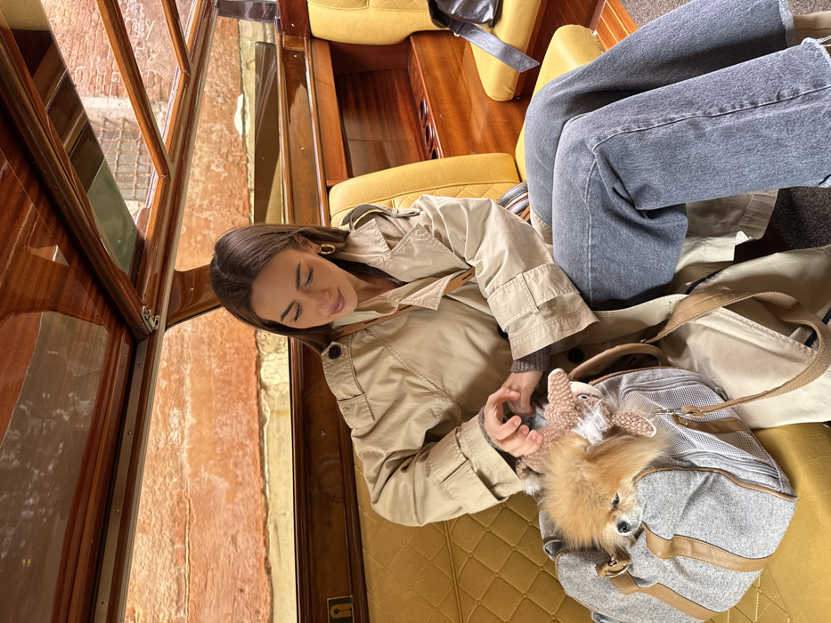A female individual is seated inside a water taxi navigating a canal in Venice, Italy. The boat's interior features polished dark wood trim, a light wood panel ceiling, and yellow quilted leather seating. Outside the boat's large windows, a narrow canal is visible, bordered by an aged, reddish-orange brick building wall. The woman, with long, dark brown hair and gold hoop earrings, is wearing a beige trench coat over a grey sweater and dark grey wide-leg jeans. Her gaze is directed downward, and her hands are positioned above a grey fabric pet carrier bag resting on the seat in front of her. A small, fluffy, light brown Pomeranian-type dog is visible within the carrier, along with a light brown stuffed animal. The woman appears to be interacting with the dog or adjusting the stuffed animal. A dark bag is partially visible on the seat to her right.