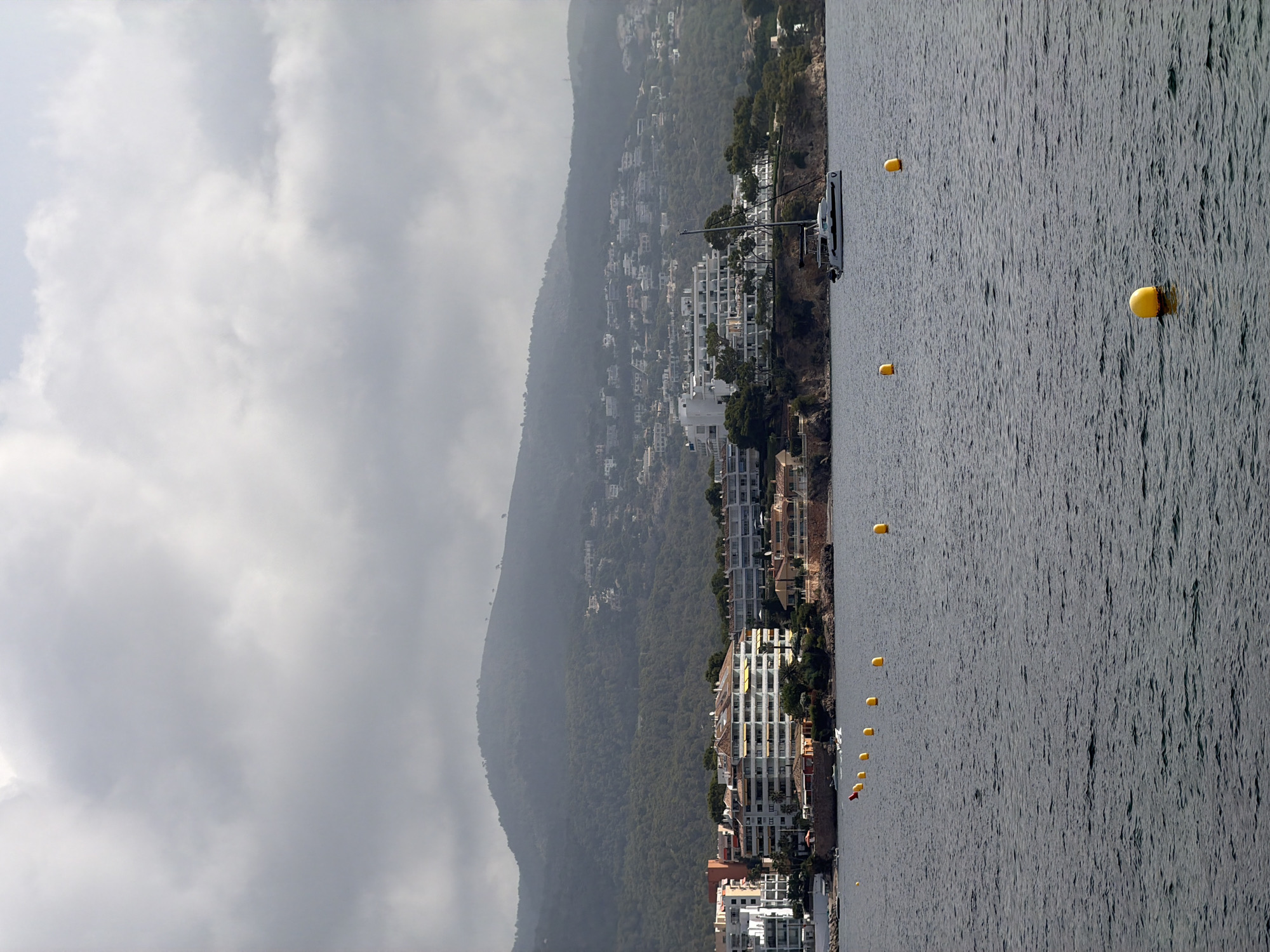 An offshore perspective captures a coastal town with a mountainous backdrop under a cloudy sky. The foreground displays a calm, dark body of water dotted with yellow buoys. In the mid-ground, a white catamaran sails on the water. The coastline features a row of multi-story buildings with white facades and yellow awnings, nestled amongst trees. Further inland, more buildings are visible on the lush green, tree-covered slopes of the mountain. The scene is located in the city of Calvià, Spain.