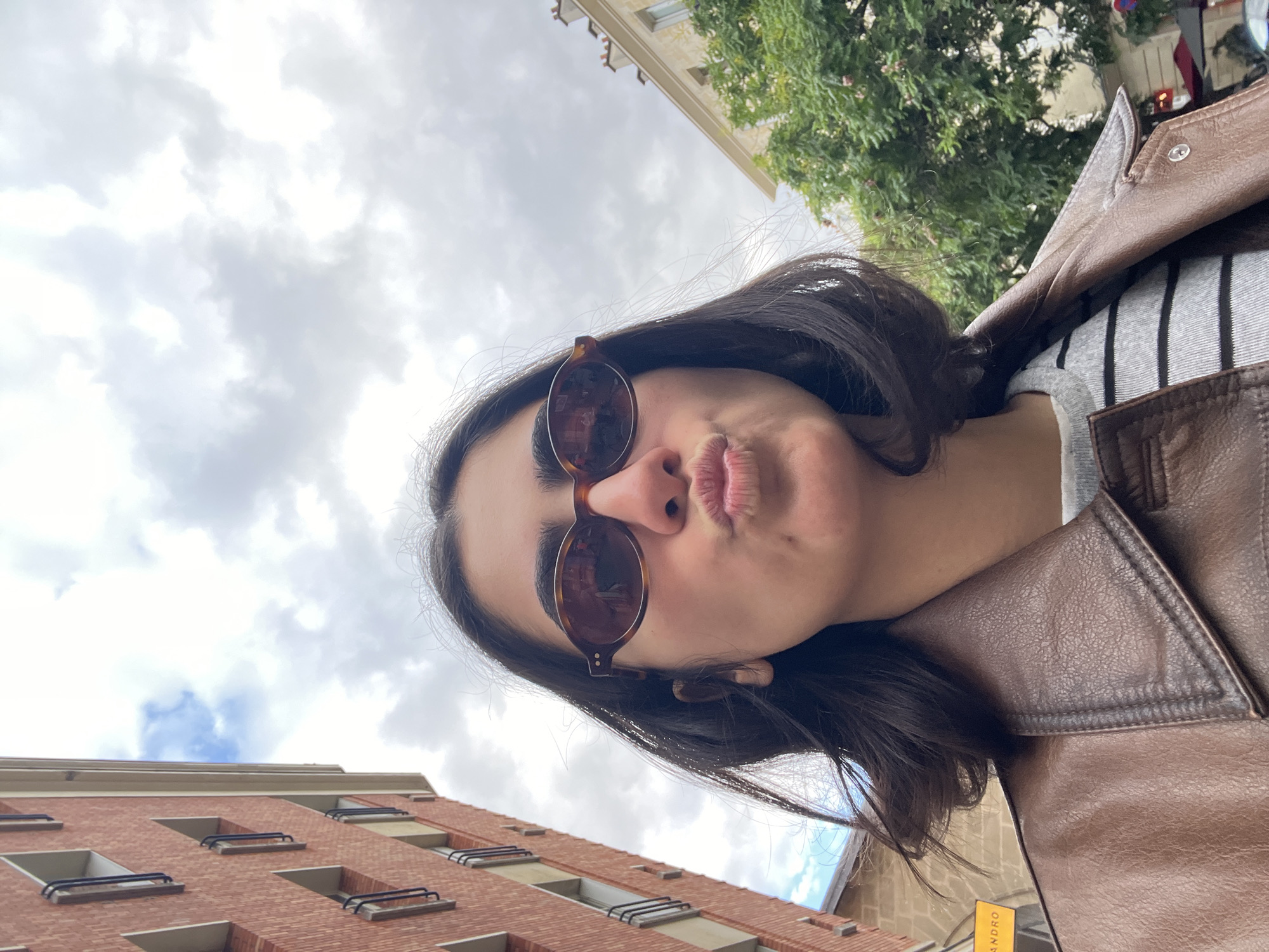 A low-angle, close-up, selfie-style photograph captures a young adult female from the chest up, positioned centrally in the frame with her head tilted slightly upward. She has dark, shoulder-length hair and dark, full eyebrows. Her lips are puckered in a distinct pouting expression. She wears oval, tortoiseshell-patterned sunglasses, reflecting light sources and parts of the surrounding environment on their lenses. Her attire consists of a brown, collared faux leather jacket worn over a grey and black horizontally striped top.
The background depicts an urban street scene in Paris, France. To the left, a multi-story building with a reddish-brown brick facade rises sharply, featuring multiple rectangular windows with white frames and black horizontal safety bars. A small patch of blue sky is visible between this building and the cloudy sky. In the lower-left corner, a partial yellow sign with black text, resembling "ANDRO" (likely part of a "SANDRO" store sign), is visible against a light-colored stone wall. To the right, an older, light-colored (yellowish-beige) stone building with visible roofline chimneys is partially obscured by a large, green-leafed tree. The sky is predominantly overcast, filled with various shades of grey and white clouds.