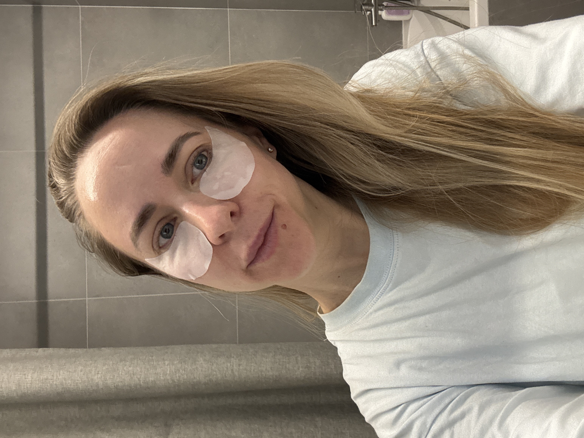 A woman with long, straight blonde hair and fair skin is captured in a close-up, slightly angled indoor shot. She has blue eyes and is looking directly at the camera with a neutral to subtly pleasant expression, hinting at a soft smile. Two translucent white under-eye patches are applied beneath her eyes, indicating a skincare routine. She is wearing a light blue, long-sleeved t-shirt, and a small, subtle stud earring is visible in her right earlobe.
The background features light grey tiled walls, typical of a bathroom setting, with a metallic fixture, possibly part of a shower or a rail, visible in the upper right. Below the tiled section, a lighter grey, textured wall covering or fabric is present. The overall lighting suggests an indoor environment. The image depicts a moment of self-care or relaxation. There is no visible text in the media file. The context for this scene is Chaikovsky, Russia.