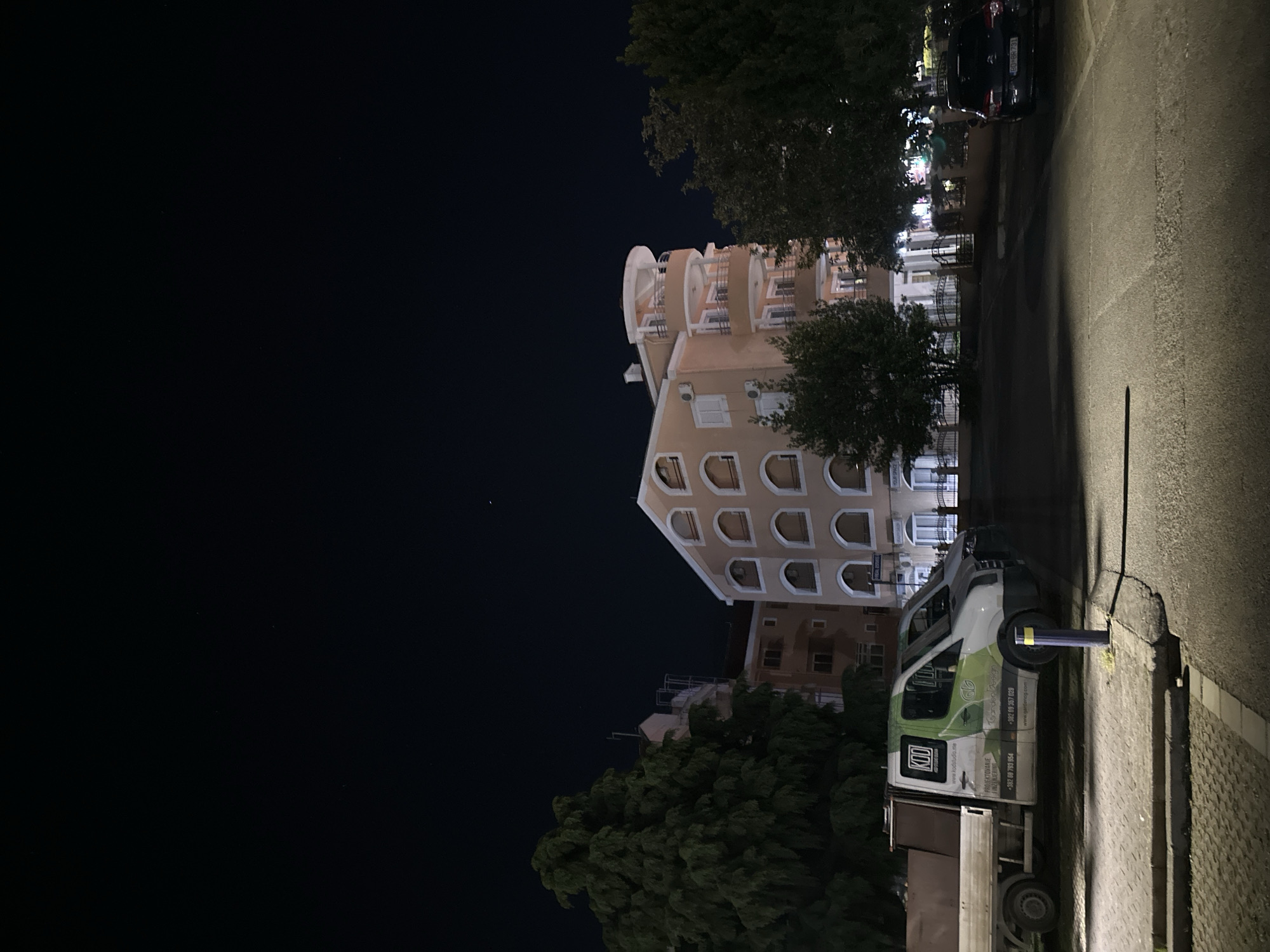 The image depicts a nighttime scene in Budva, Montenegro, showing a light-colored building with distinctive arched windows and a rounded turret. A white and green truck with visible branding including "KOD" and "Garden Design" is parked along the road in front of the building. The asphalt road is illuminated by streetlights, and a dark-colored car is parked further up the road. Trees are interspersed between the building and the road. The sky is dark with a few visible stars. Some of the signage on the van's door is visible.