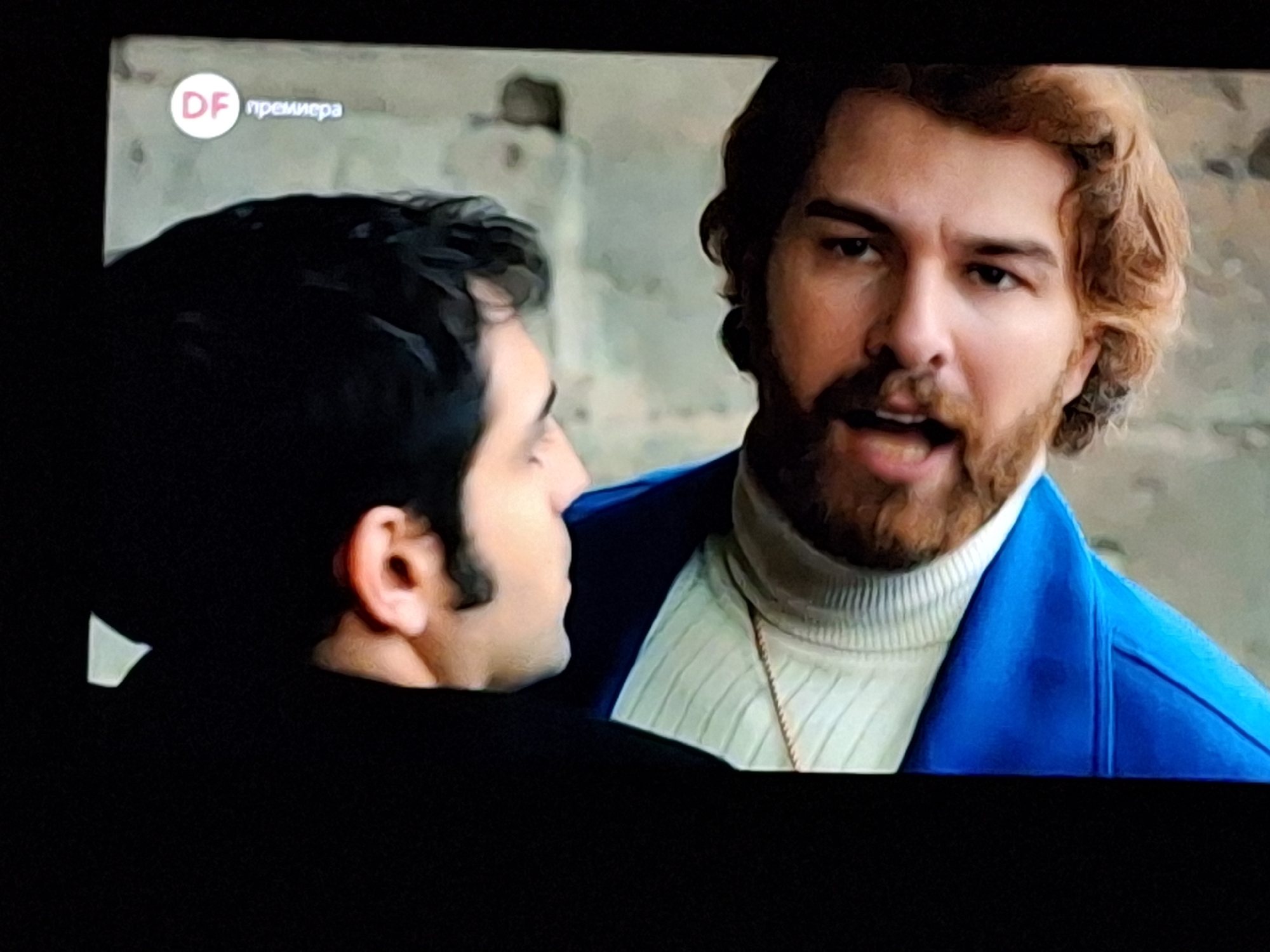 The image shows two men in a close interaction. One man has dark hair and is wearing a black shirt, with only the side of his face and ear visible. The other man, facing the viewer, has reddish-brown hair and a beard. He is wearing a white turtleneck sweater underneath a bright blue blazer. The scene is likely from a television show, indicated by the "DF премьера" logo in the upper left corner of the image. The background is blurred, but there appears to be a concrete wall. Based on the location in the city of Zverino, Bulgaria, the scene could be a segment from a television series or movie. The men seem to be in conversation, with the man in the blue blazer possibly speaking.