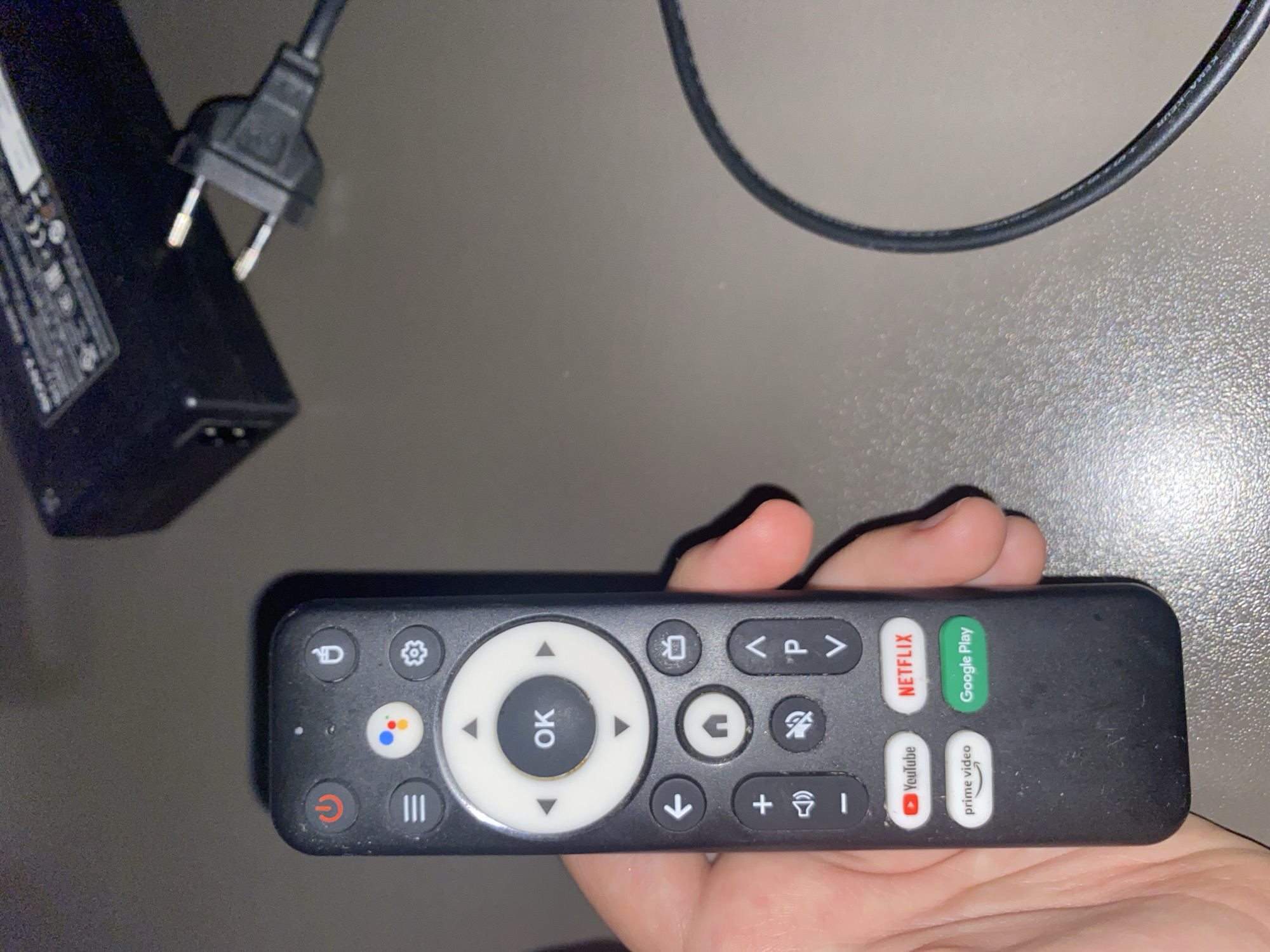 The media file shows a close-up, overhead shot of a hand holding a black TV remote control. The remote has a central navigation ring with an "OK" button in the middle. Above the navigation ring are buttons for settings and home. Below the navigation ring are buttons for back and recent apps. To the right of the navigation ring are buttons for "V < P >" and mute. Below that are volume up/down and input select buttons. There are also dedicated app buttons for Netflix, Google Play, YouTube, and Prime Video. The background is a plain, light gray surface, possibly a table, with a blurry black power adapter and cable visible in the upper left corner. The scene appears to be indoors, with neutral lighting and no indication of the time of day or weather. The focus is on the remote control and the hand holding it, with no discernible emotions or specific activities other than the act of holding the remote. Visible text on the remote includes "OK", "Netflix", "Google Play", "YouTube", and "prime video". The power adapter also has some legible text, but it is too blurry to decipher. The location context provided is Tolyatti, Russia, but there are no visual cues within the image to confirm this.