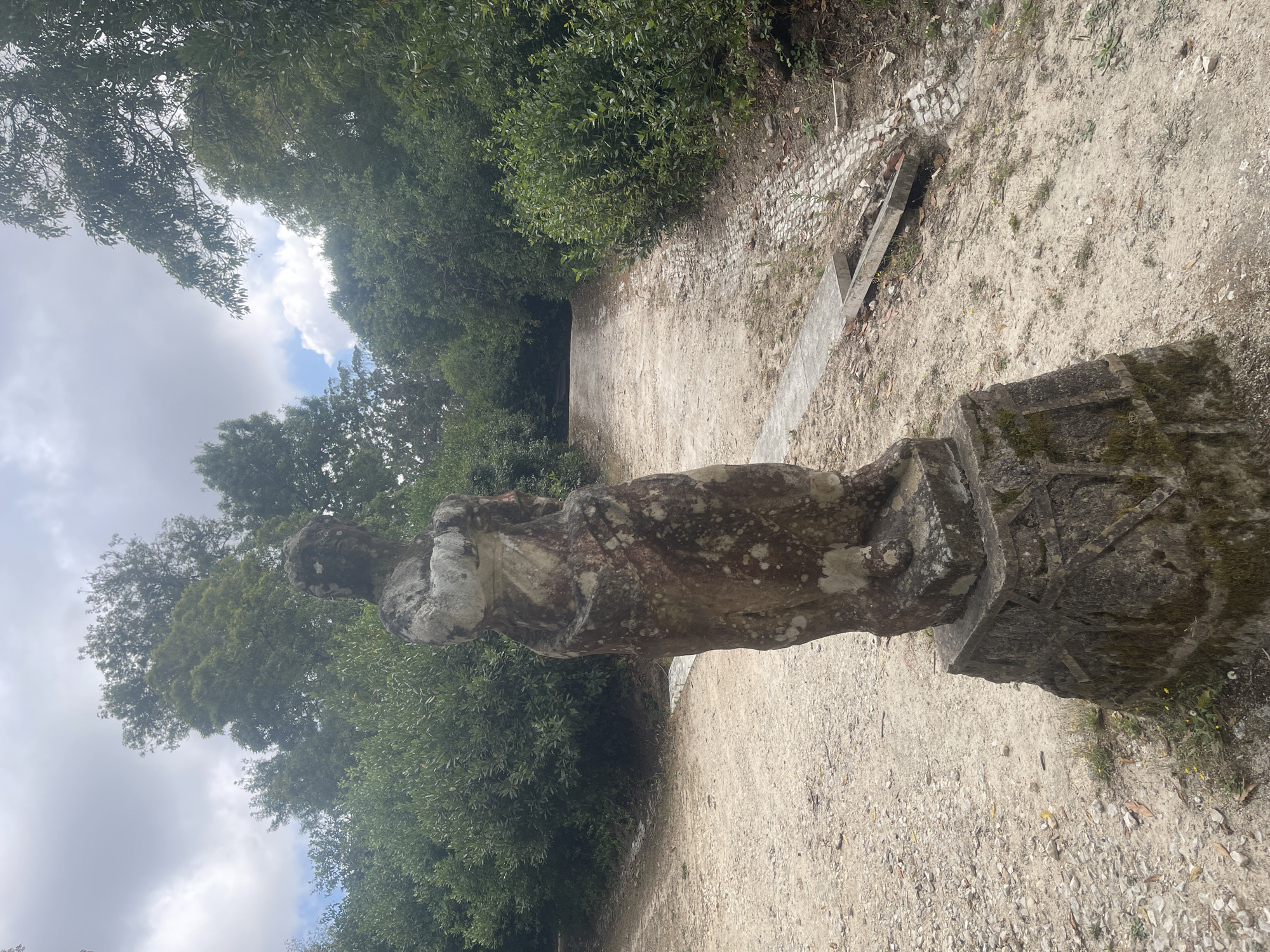 An aged, weathered stone statue stands on a multi-tiered pedestal in the foreground of an outdoor scene. The statue depicts a figure draped in what appears to be a robe, with its arms crossed. The stone is textured and discolored with moss and lichen, suggesting it is old. The pedestal is also made of stone and has some decorative carvings.
Behind the statue, a dirt path winds its way uphill, flanked by dense green foliage on both sides. The path is uneven and appears to be made of gravel and earth, with some exposed stones and a cobblestone section in the middle. There is a concrete channel or drain running alongside a portion of the path.
The sky above is partly cloudy with patches of blue sky peeking through. The surrounding trees are lush and green, suggesting a temperate climate. The overall impression is one of an overgrown, historical park or garden.
This statue is located in the gardens of the Mafra National Palace, in Mafra, Portugal. The palace is a UNESCO World Heritage site, and its expansive gardens are a popular attraction.