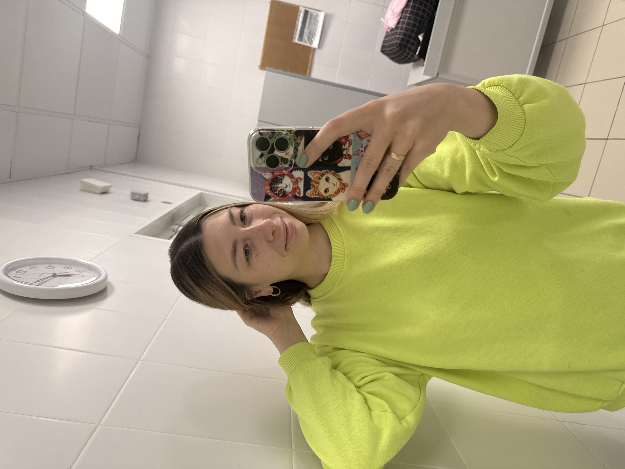 A young woman is holding a smartphone to take a selfie. She is wearing a neon green sweatshirt. Her left hand is resting on her hair. She has fair skin and blond hair. Her nails are painted light green. She is wearing a ring on her finger. The smartphone has a case with cartoon cats. A clock is visible on the tiled surface below. The room has white tiled walls. In the background, there is a mirror and a bulletin board. The location appears to be a restroom, possibly in Moscow, Russia given the aesthetic similarities to public spaces there.