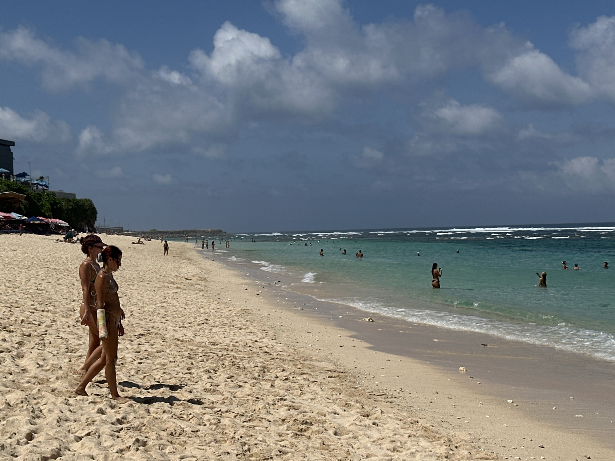 Two women walk on the sandy beach of Pandawa Beach in Kuta Selatan, Indonesia. One of the women has a cast on her arm. Other people are swimming in the ocean. The weather is sunny with partly cloudy skies. Along the back of the beach are trees and some buildings.