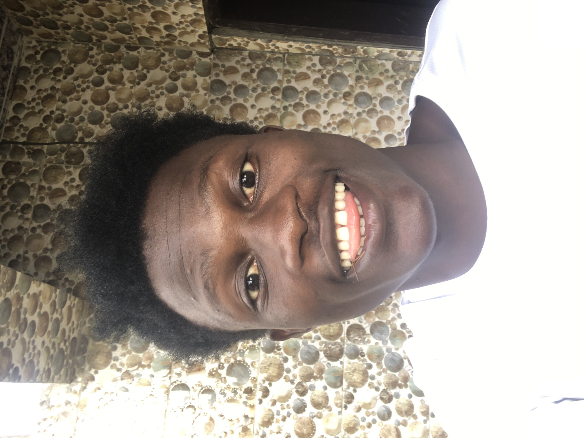 A young Black male is pictured in a close-up, head-and-shoulders selfie. He has short, dark, curly hair and brown skin. He is wearing a white t-shirt. He is smiling with his mouth open, showing his teeth and tongue. A small twig is visible between his teeth. Behind him, the wall is covered in small, circular tiles with a mosaic pattern in shades of brown, beige, and grey. The lighting suggests he is outdoors, possibly near an entrance, given the dark doorway visible on the right side of the frame. The background pattern is consistent with decorative tiling often found in residential or commercial exteriors in areas like Ajah, Nigeria.