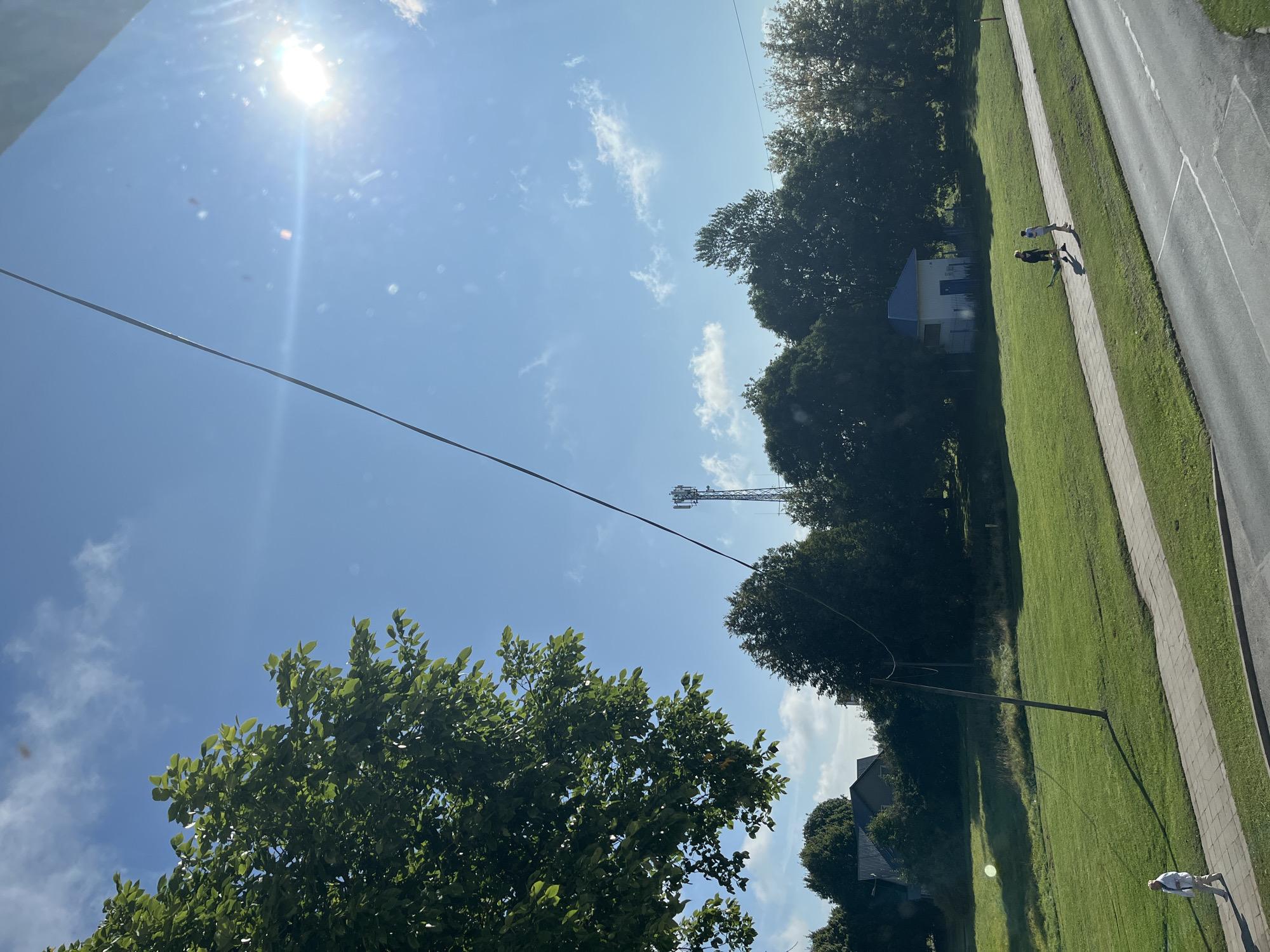The image depicts an outdoor scene in Kohtla-Järve linn, Estonia, under a bright, sunny sky with scattered white clouds. A power line stretches diagonally across the frame from the upper left to the lower right. In the foreground, a paved sidewalk runs parallel to a road. A person, dressed in light-colored clothing, walks along the sidewalk. To the left of the sidewalk, a well-maintained green lawn is visible, with trees and houses in the background. Further along the sidewalk, two individuals are walking away from the camera. Behind them, a small blue and white building with a dark blue door is situated among trees. In the distance, a cellular tower stands prominently against the sky. The sun is bright and positioned in the upper right quadrant of the image, casting shadows across the lawn. The overall scene suggests a clear, warm day in a semi-urban or suburban environment.