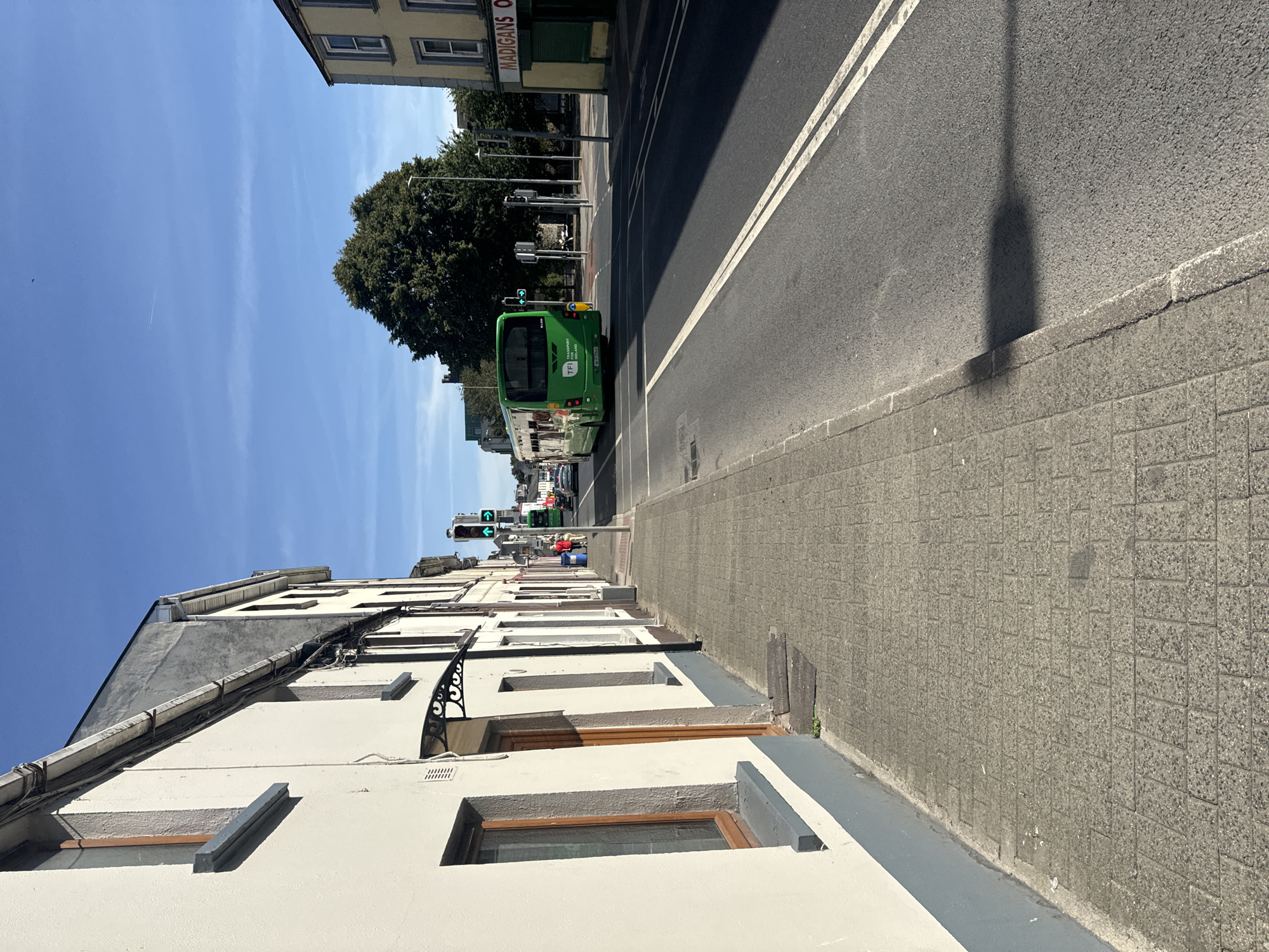This is a street-level, eye-level, vertical shot of a street scene in Waterford, Ireland. The image captures a bright, sunny day with a clear blue sky. The left side of the frame features the side of a cream-colored building with a gray base, featuring windows with brown frames and a small awning over a doorway. The sidewalk, paved with gray rectangular tiles, runs along the building. On the right side of the frame, the street is asphalt with double white lines. A green bus with "Transport for Ireland" branding is in the center of the road, moving away from the camera. Traffic lights, showing green arrows, are visible ahead. Further down the street, buildings line the right side, one with a sign reading "MADIGANS O". A large tree with dark green foliage stands near the center-right of the image. A few people are visible on the sidewalk in the distance, and a car is seen on the road. The overall scene depicts a typical urban street during daylight hours.