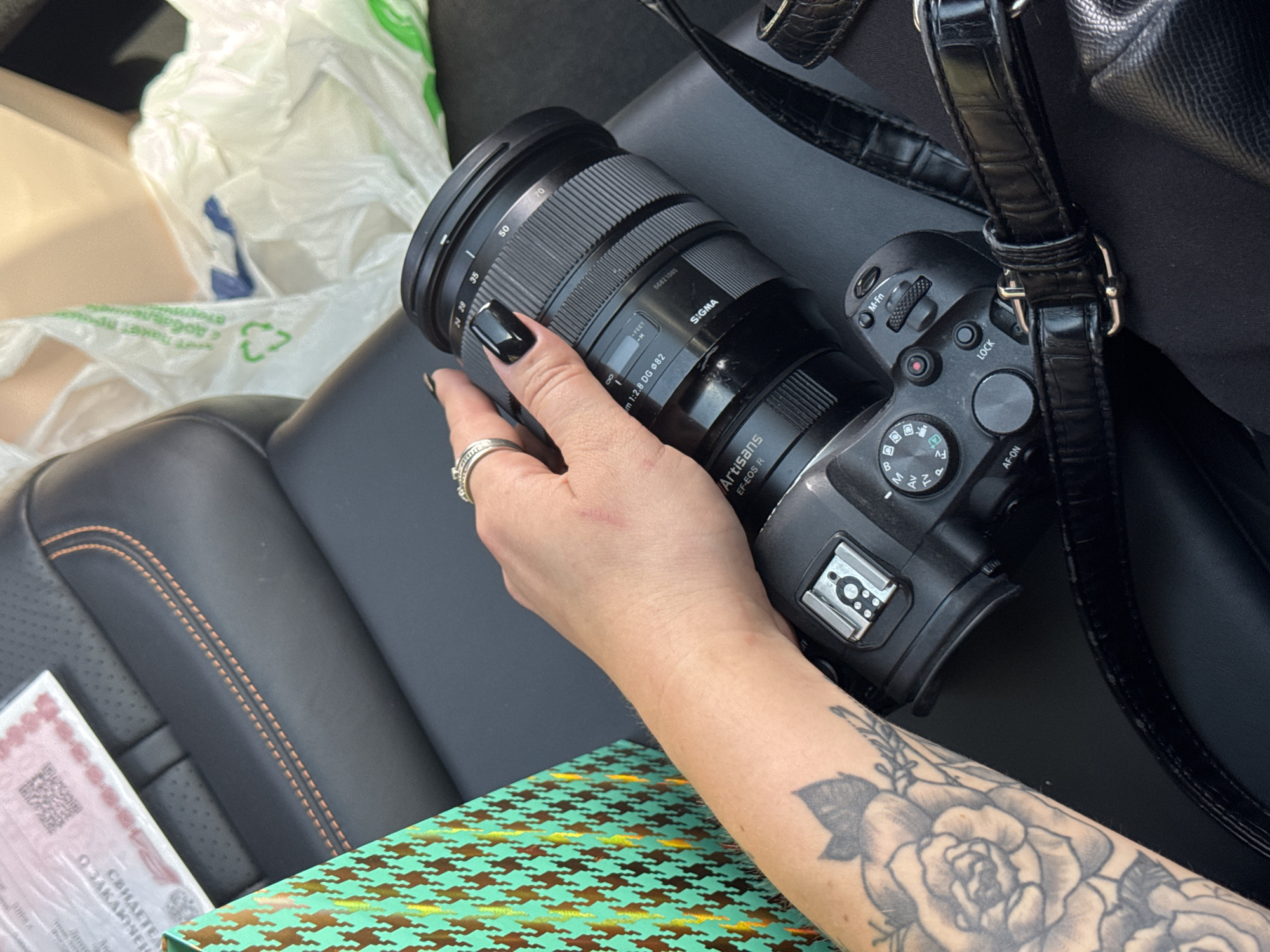 An individual's left arm, adorned with a rose tattoo and rings, is visible as they hold a Sigma camera with a lens attached. The camera is resting on a black leather surface, possibly a car seat, with orange stitching along the edge. A patterned gift bag and crumpled plastic bags are also present in the background, suggesting an indoor or vehicle setting. The scene is lit by natural light.