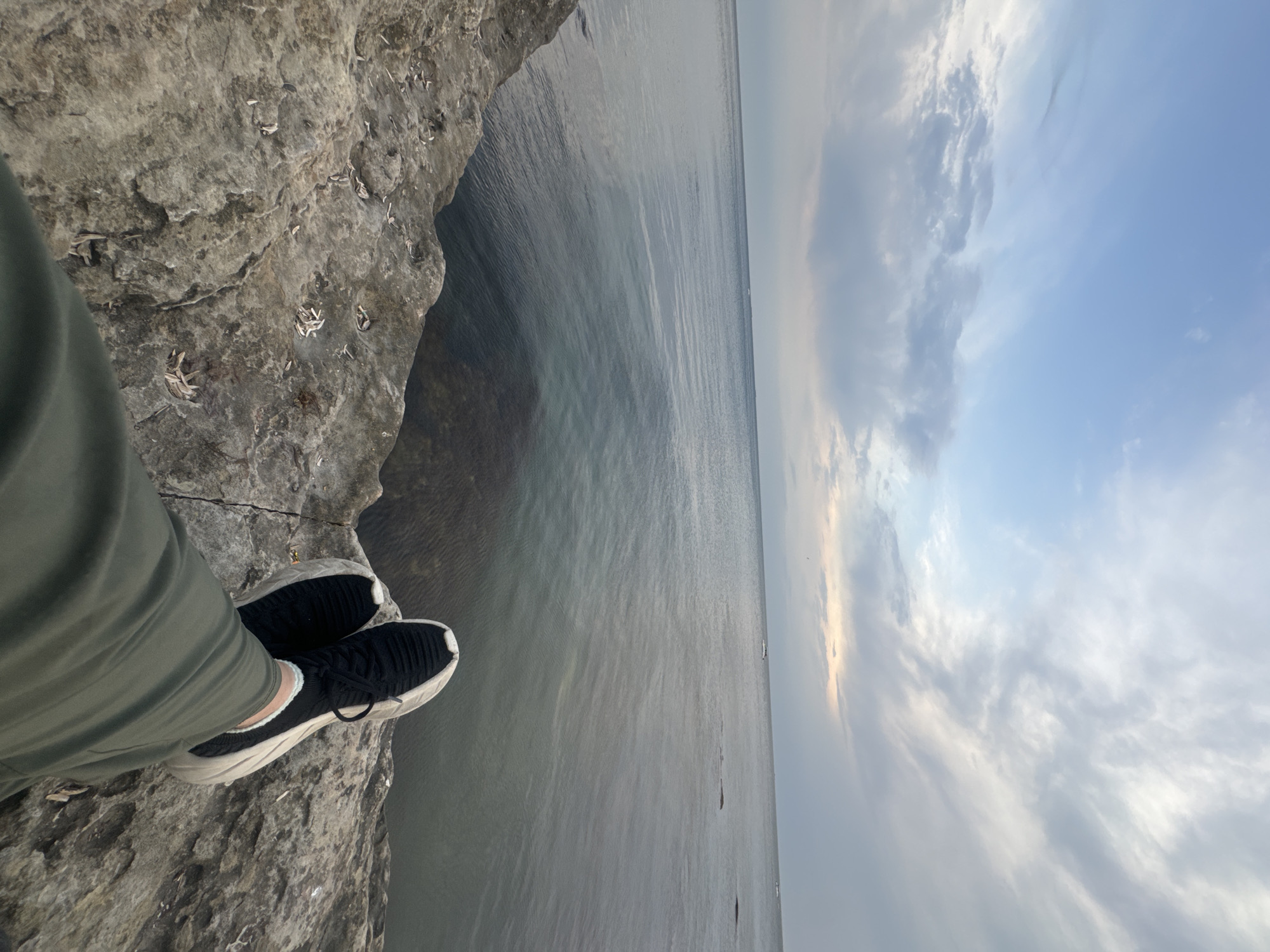 The image captures a person's legs and feet, clad in olive green pants and black athletic shoes, resting on a rocky, uneven shoreline. The perspective is from a low angle, looking out towards a calm, greyish-blue body of water that extends to the horizon. The sky above is a mix of light blue and scattered grey clouds, with a faint hint of pinkish light near the horizon, suggesting either sunrise or sunset. The rocky area in the foreground is textured and appears dry, with small debris scattered across its surface. In the distance, the water is relatively still, with subtle ripples. The overall scene is serene and suggests a moment of quiet contemplation by the water. While the prompt mentions the city of Aktau, Kazakhstan, there are no specific visual cues in the image that definitively place it there.